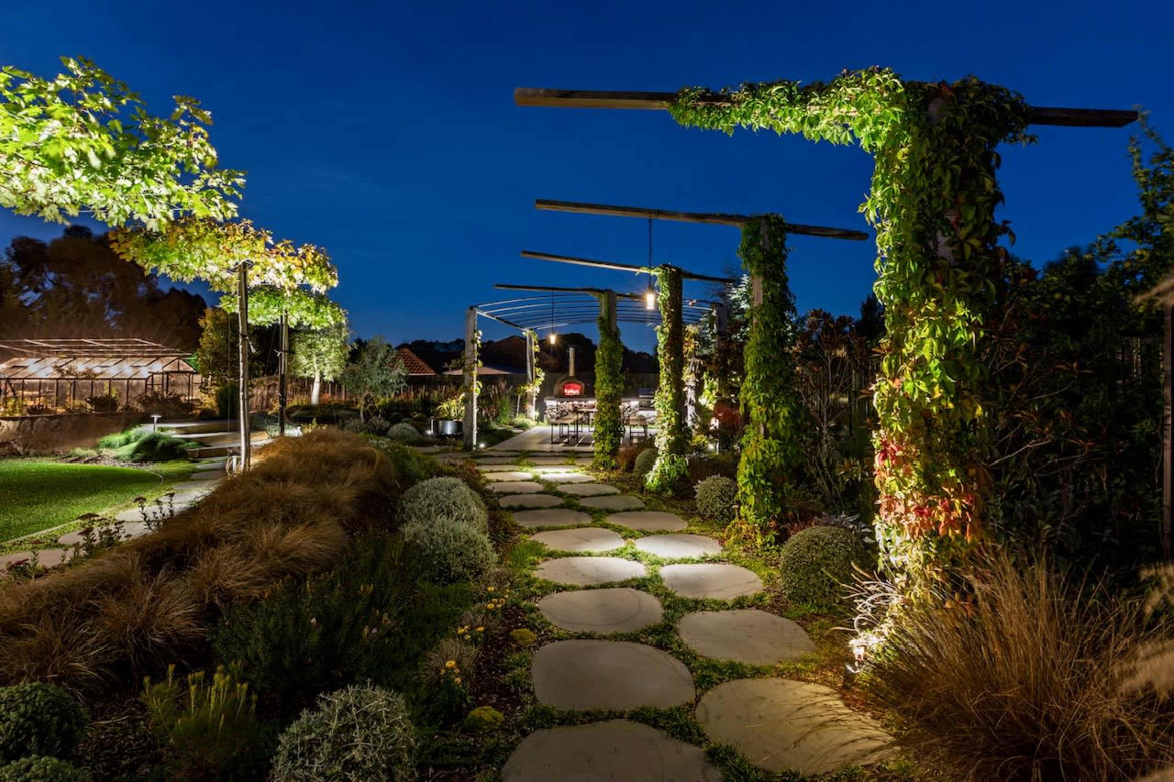 A well-lit garden path lined with stone slabs and surrounded by greenery and climbing plants under a twilight sky.