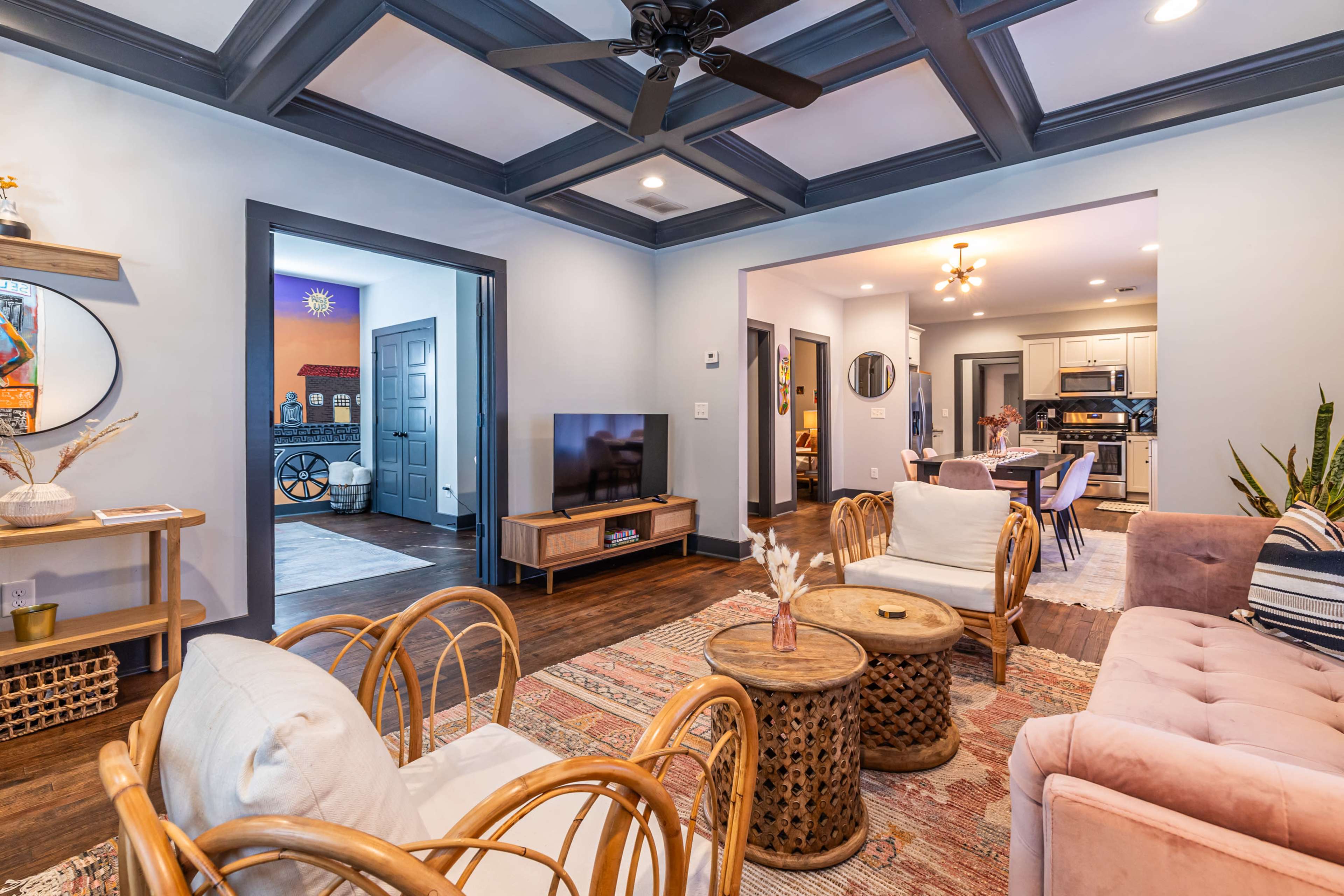 The image shows a living room with a coffered ceiling, wooden floor, and a mix of seating arrangements, including chairs and a sofa, leading to an adjacent dining area.