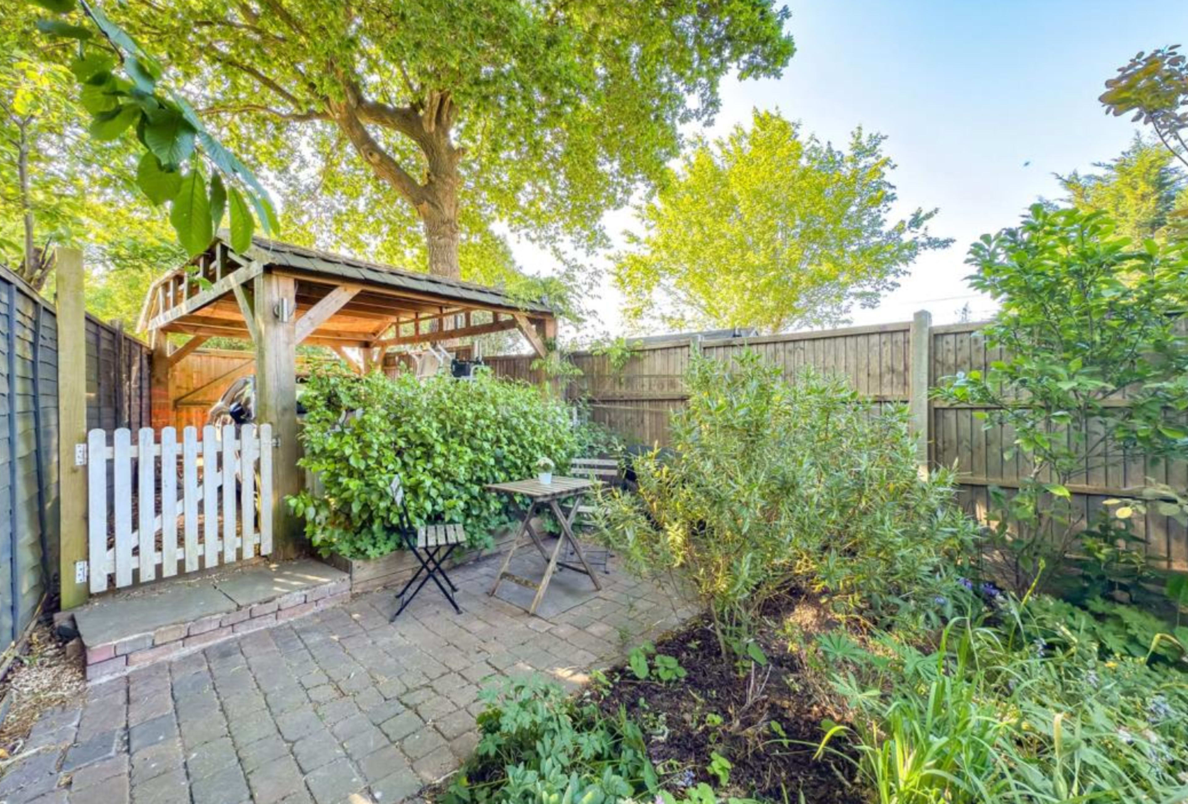 The image shows a small garden area with a wooden shed, a white picket fence, and seating surrounded by greenery.