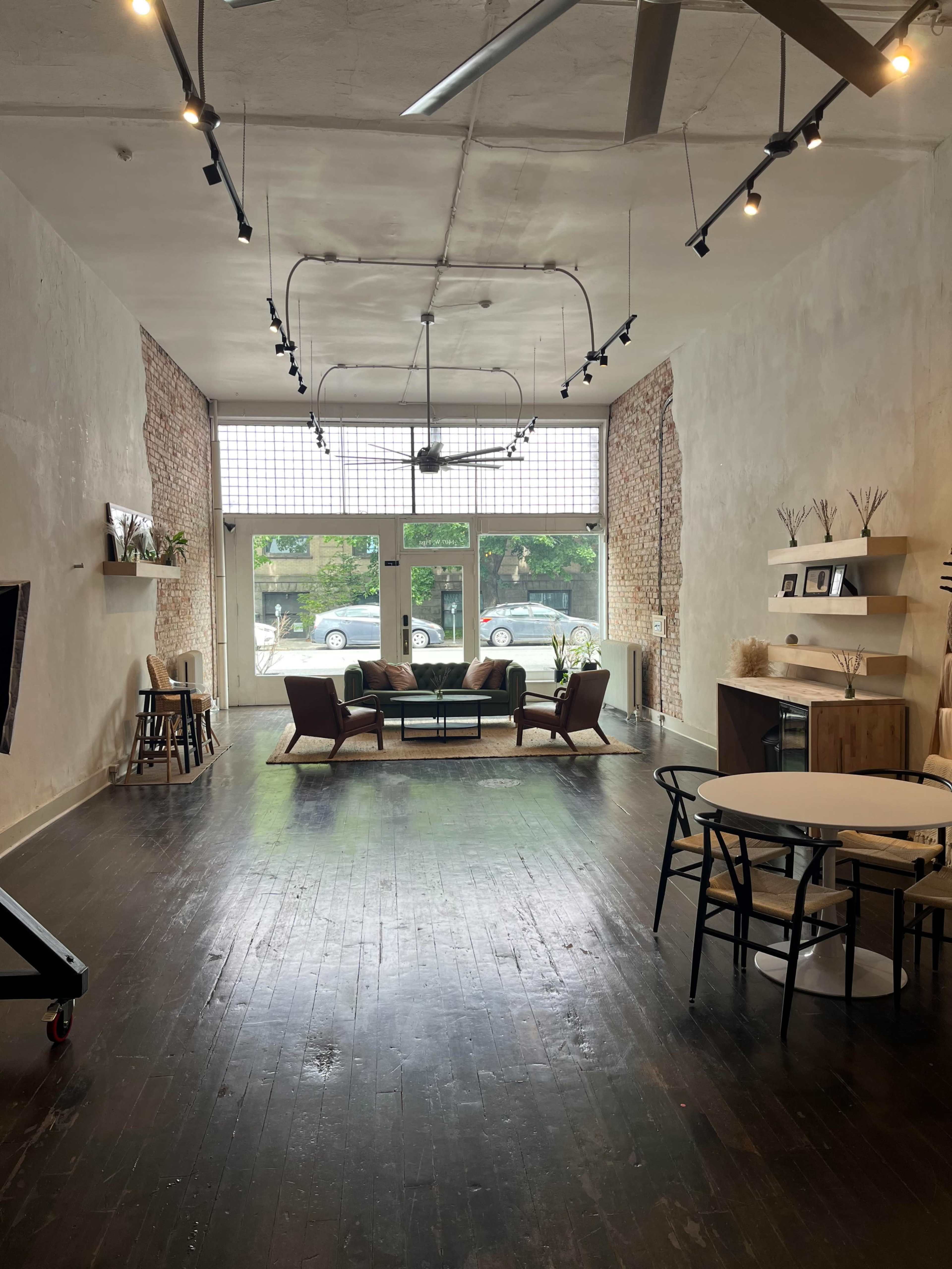 The interior of a spacious, lightly decorated room features wooden floors, large windows, and a seating area with a couch and chairs.