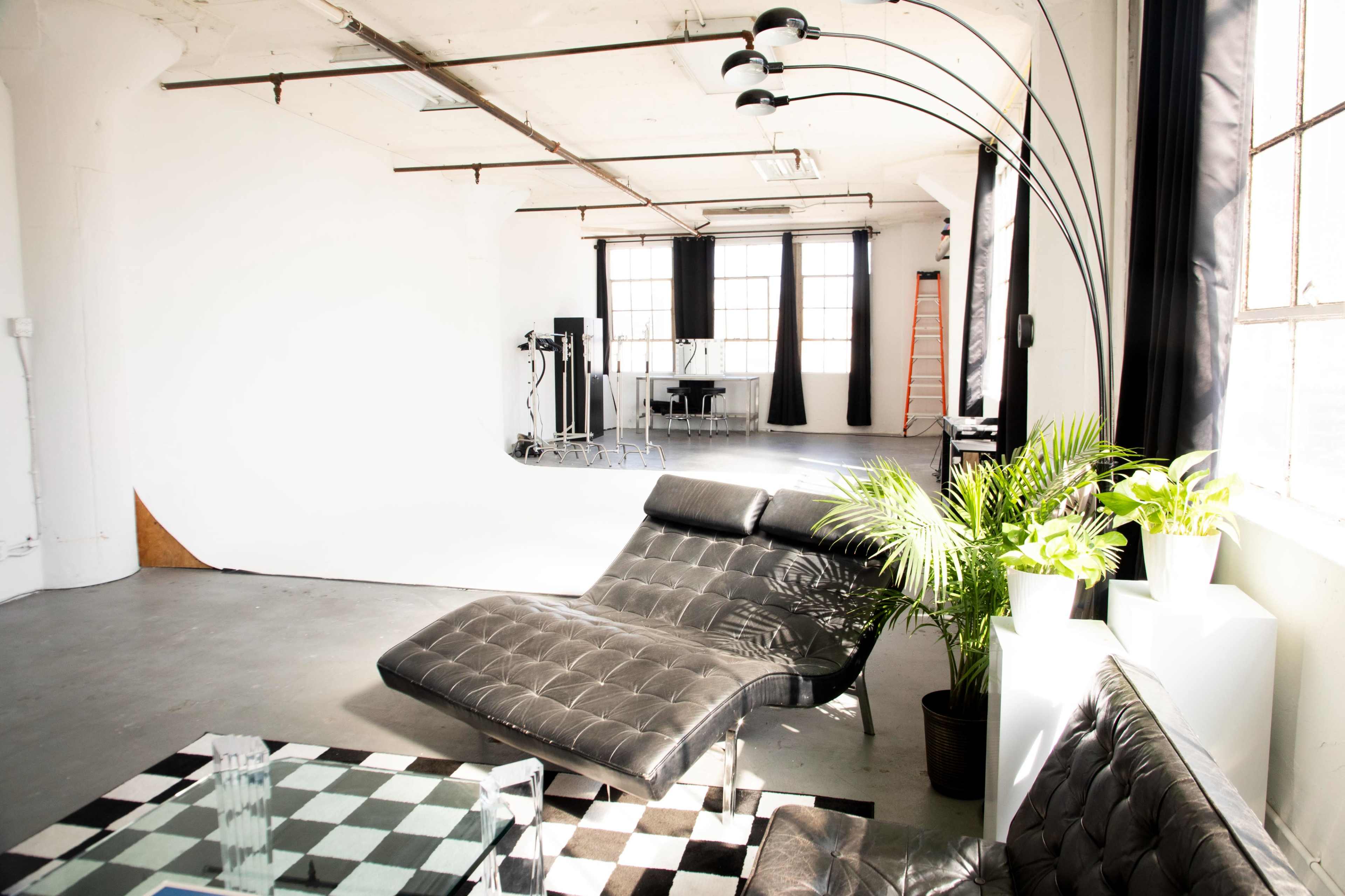 A modern studio space featuring a black lounge chair, a checkered coffee table, and large windows with curtains.
