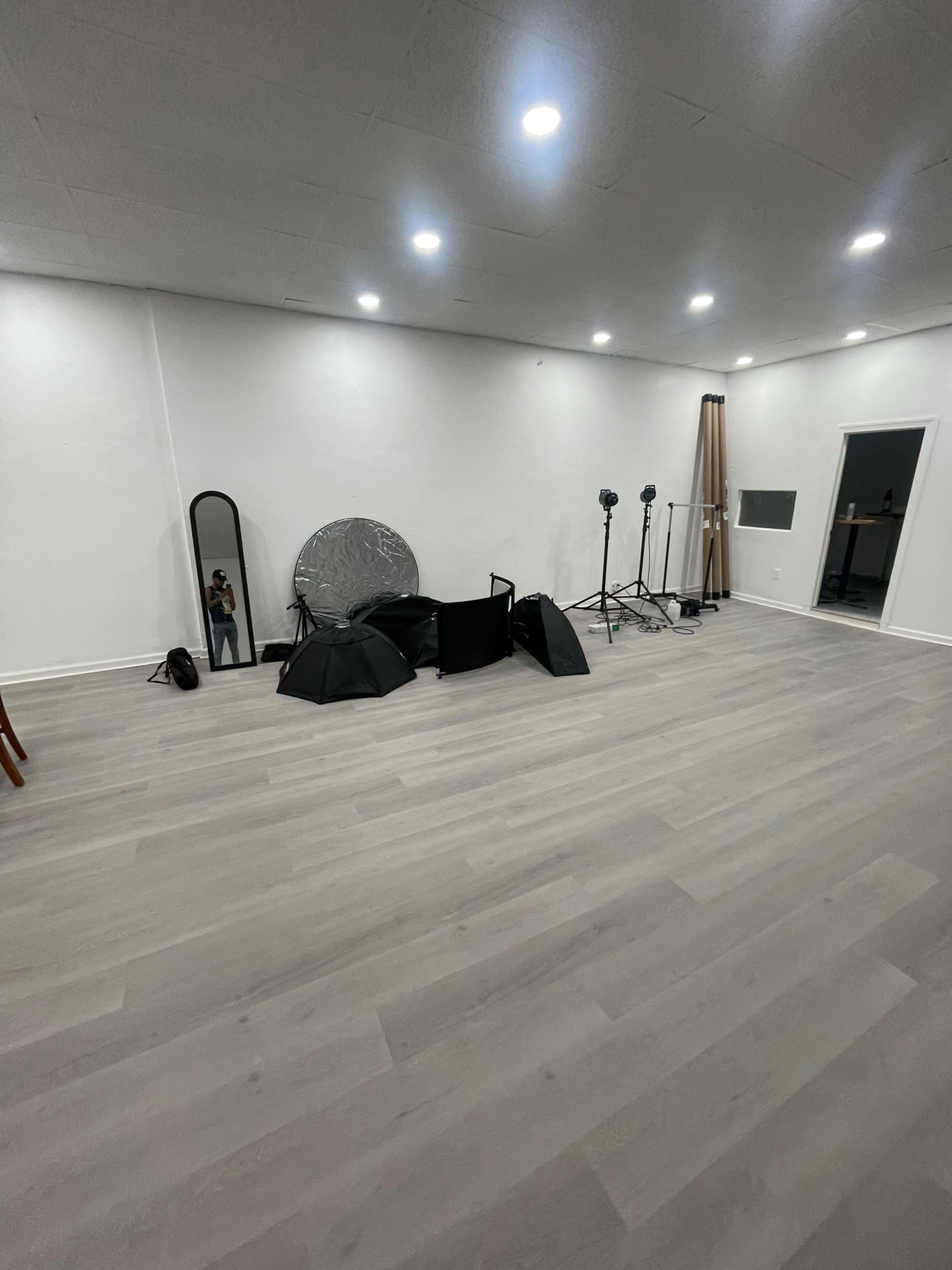 A spacious, well-lit room with a wooden floor, several photography equipment items on the ground, and a mirror against the wall.