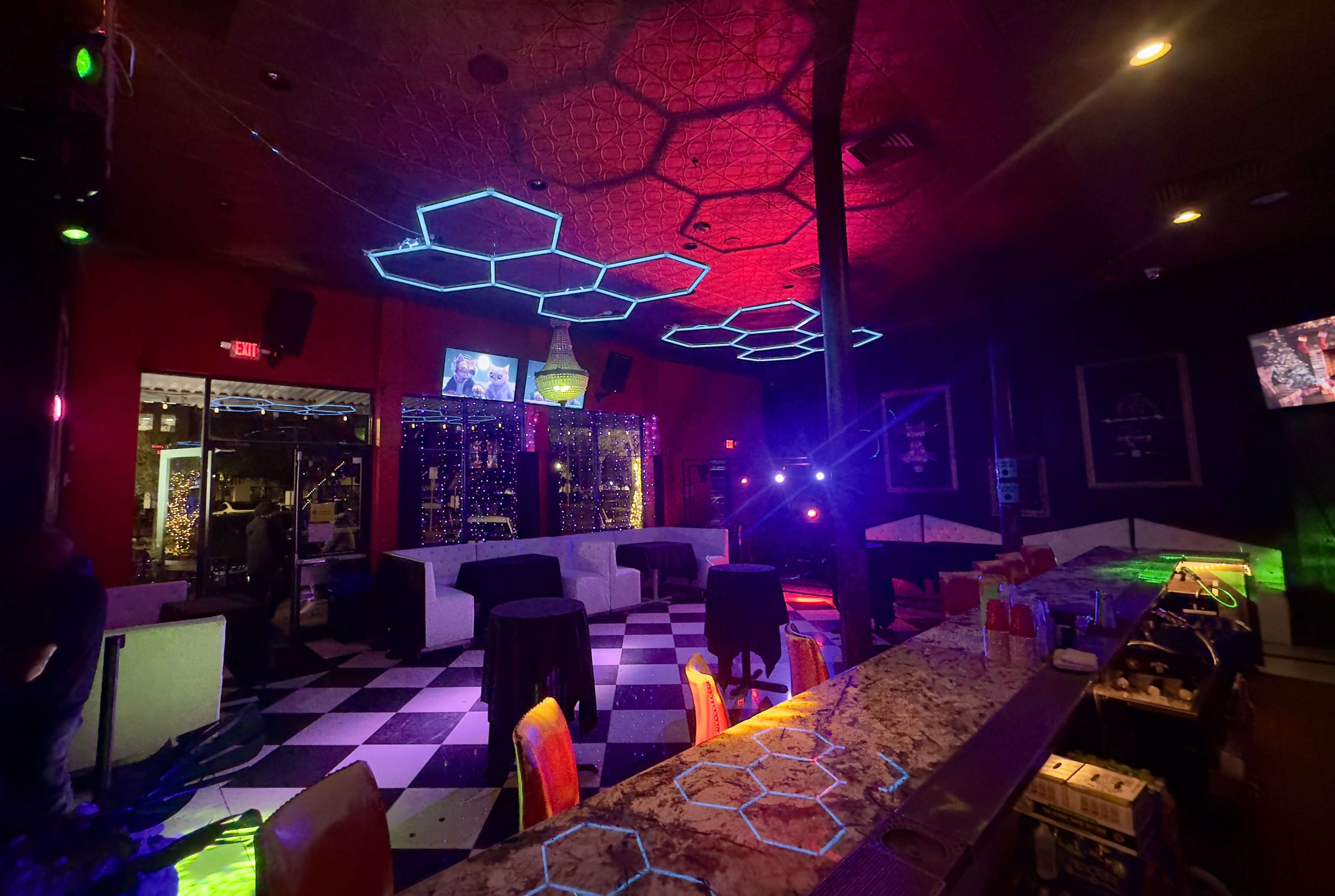 The image shows a dimly lit bar with a checkered floor, illuminated by colorful lights and geometric patterns on the ceiling.