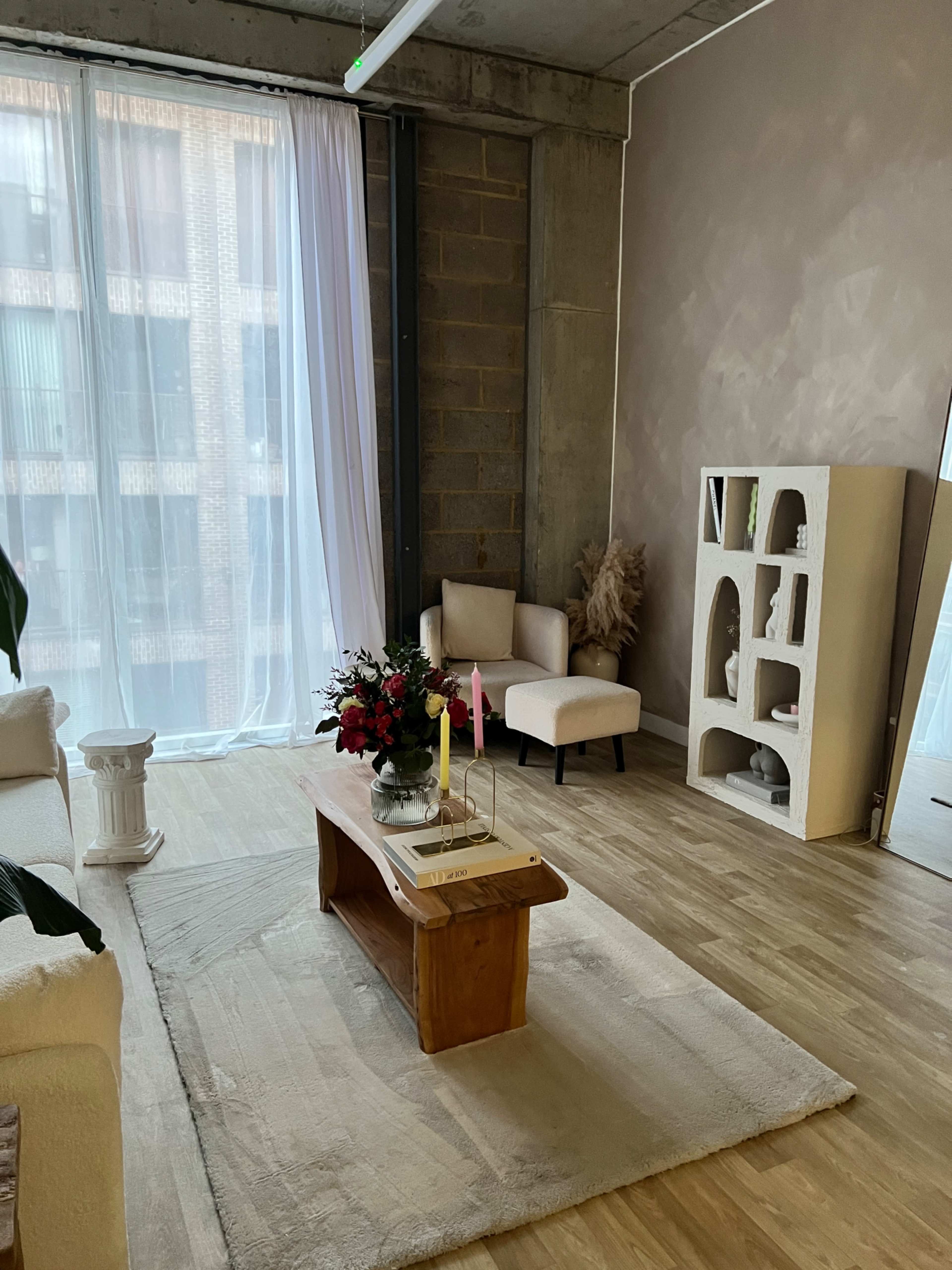 The room features a light-colored wooden floor, a large window draped in white fabric, and a coffee table adorned with flowers and candles, alongside a chair and minimalist shelving unit.