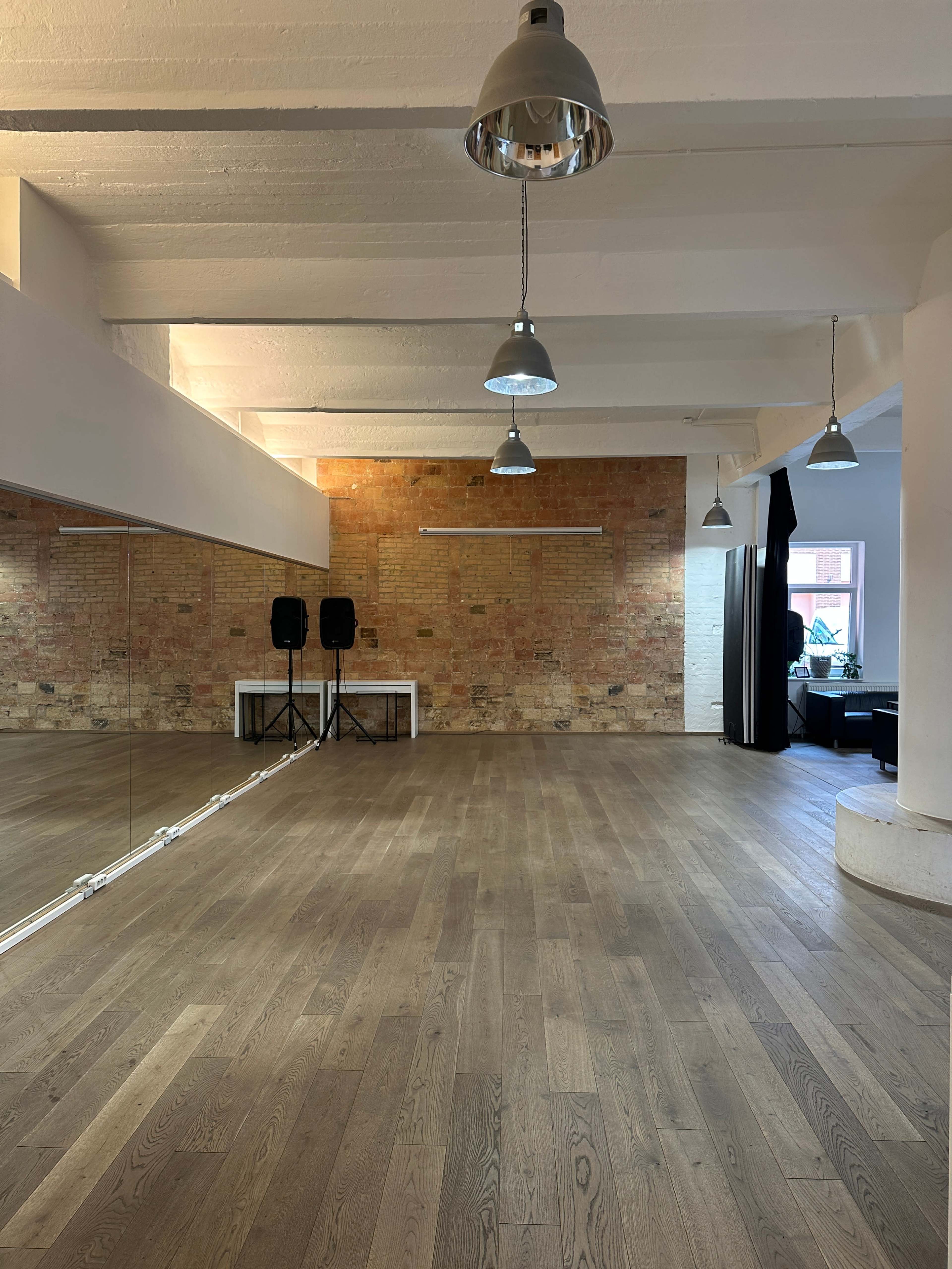 The image shows a spacious, well-lit room with wooden flooring, exposed brick walls, and a large mirror along one side.