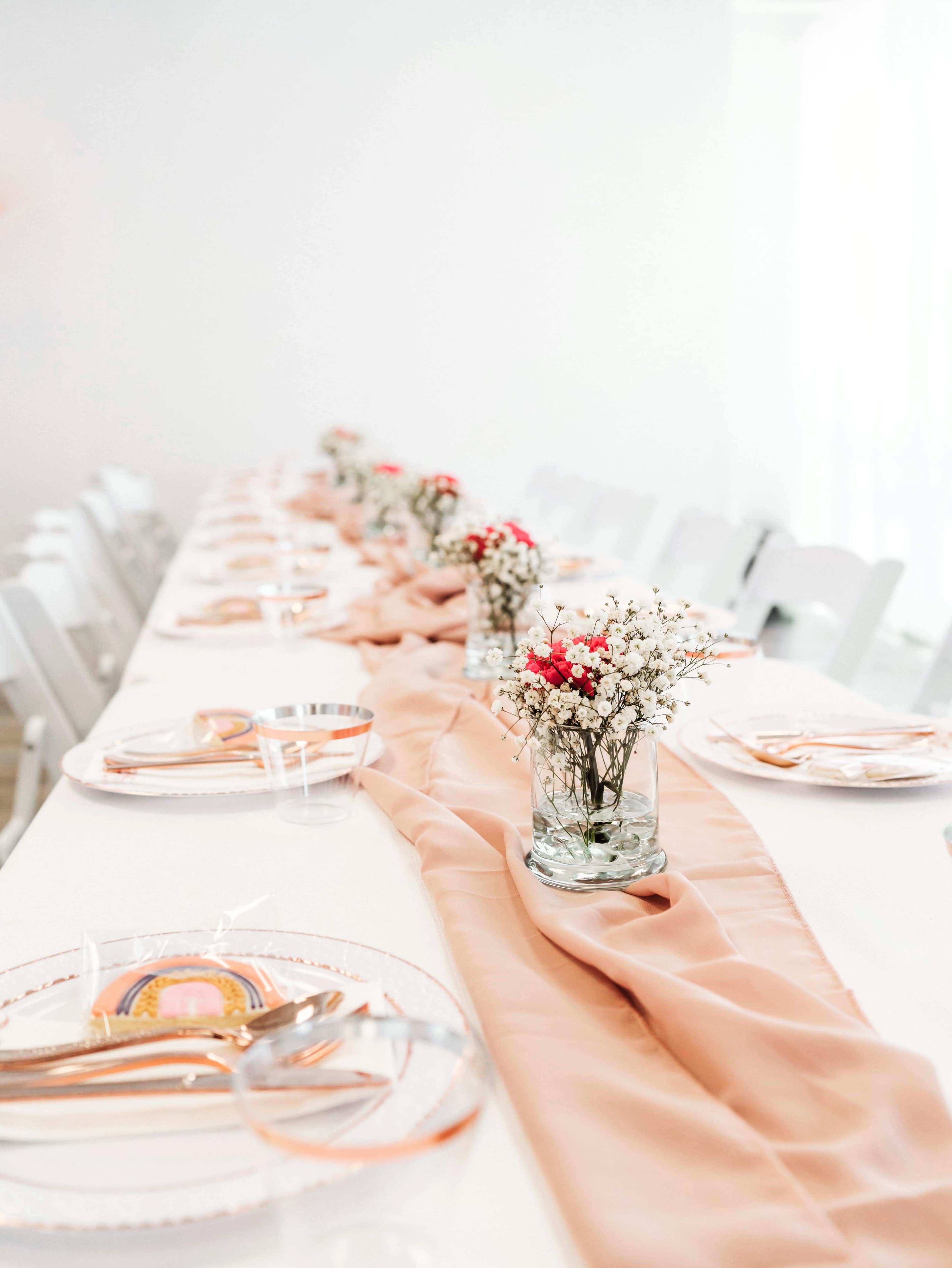 A long dining table is set with clear plates, gold utensils, and a light peach table runner, adorned with small floral centerpieces in glass vases.