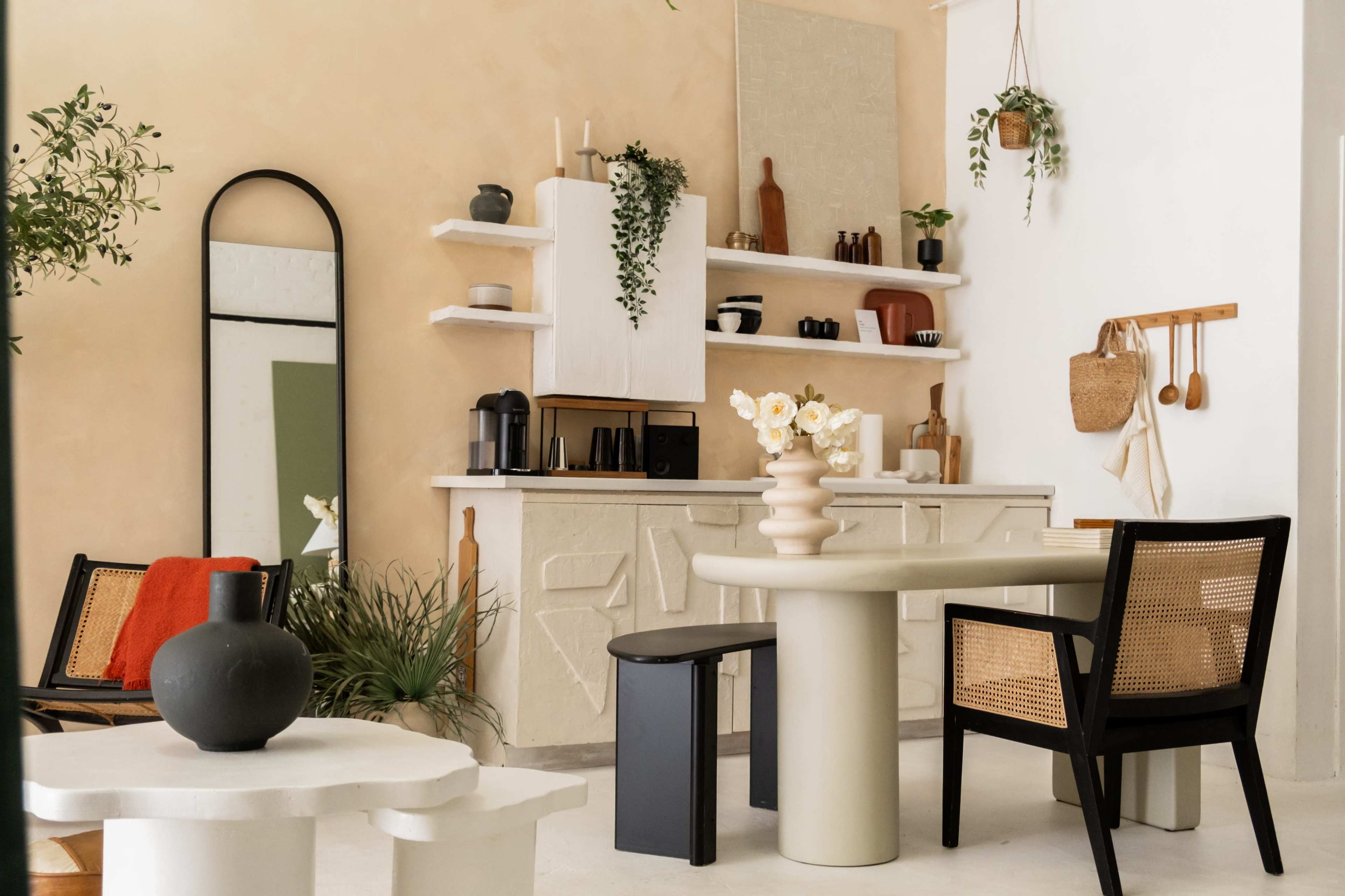 The image shows a modern, minimalist living space with a round dining table, a black chair, and a white kitchen area featuring decorative shelves and various plants.