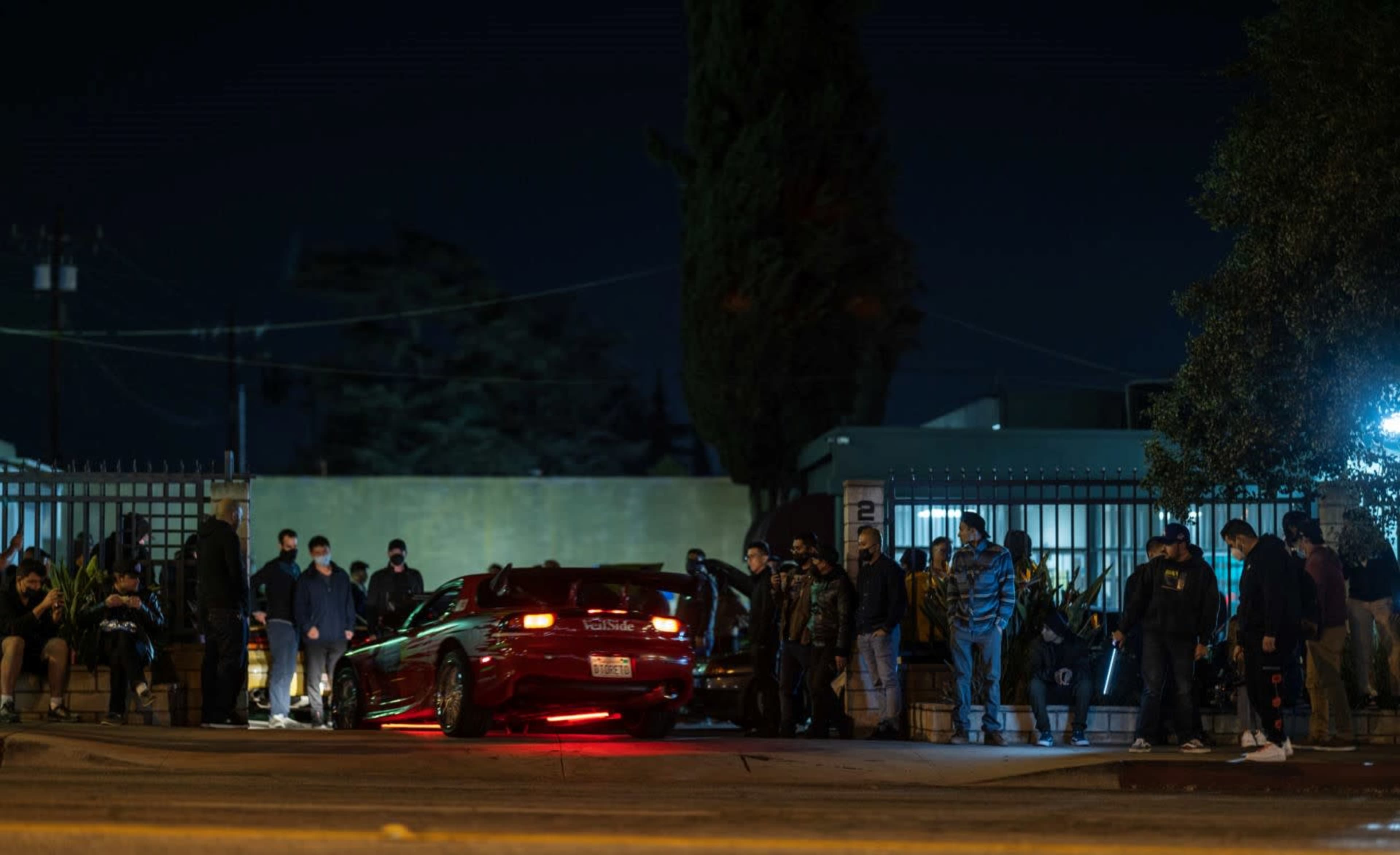 A group of people gathers outside at night near a parked car with illuminated underglow lights.
