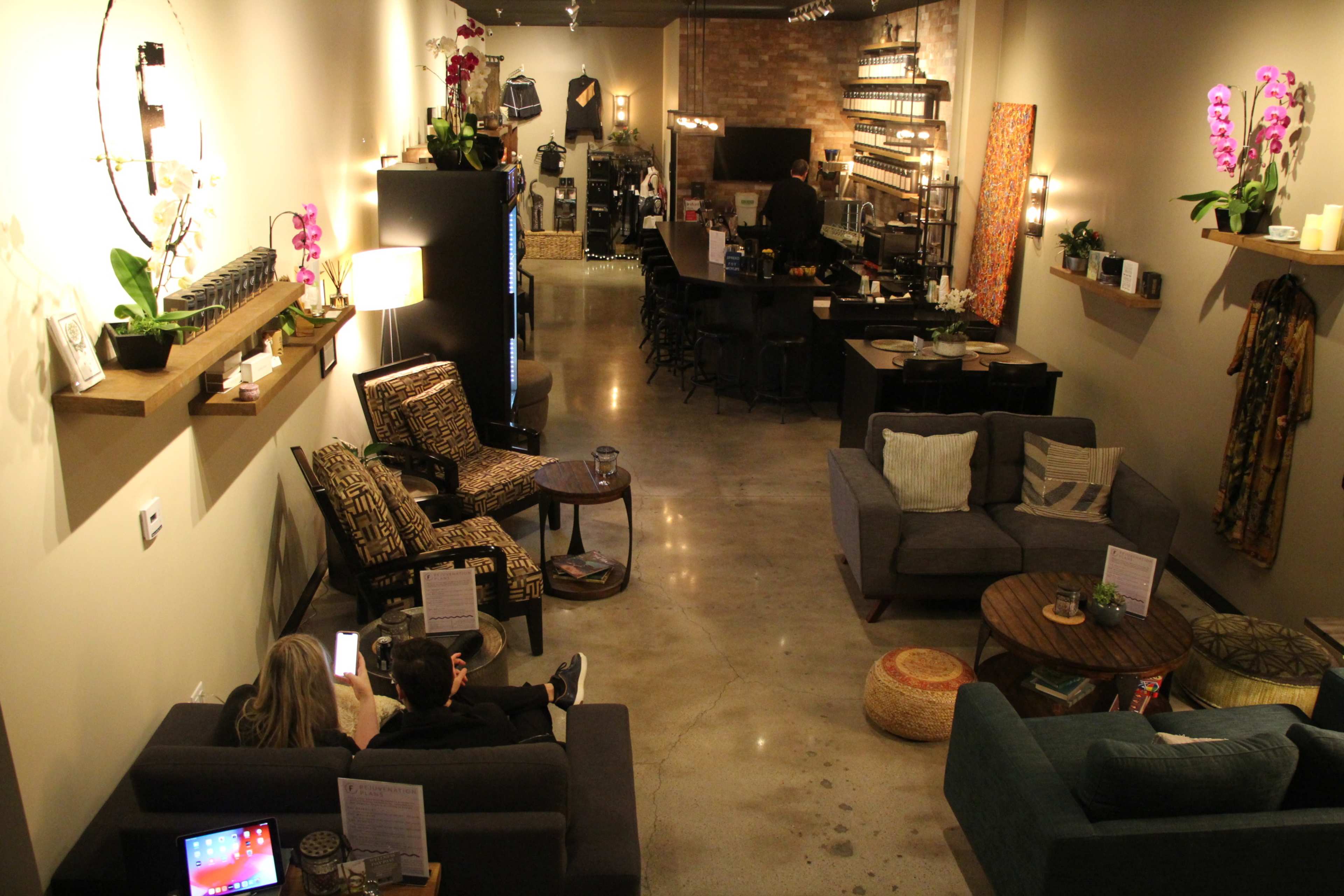 The image shows a stylish café interior with various seating arrangements, including sofas and chairs, along with decorative shelves and a bar area in the background.