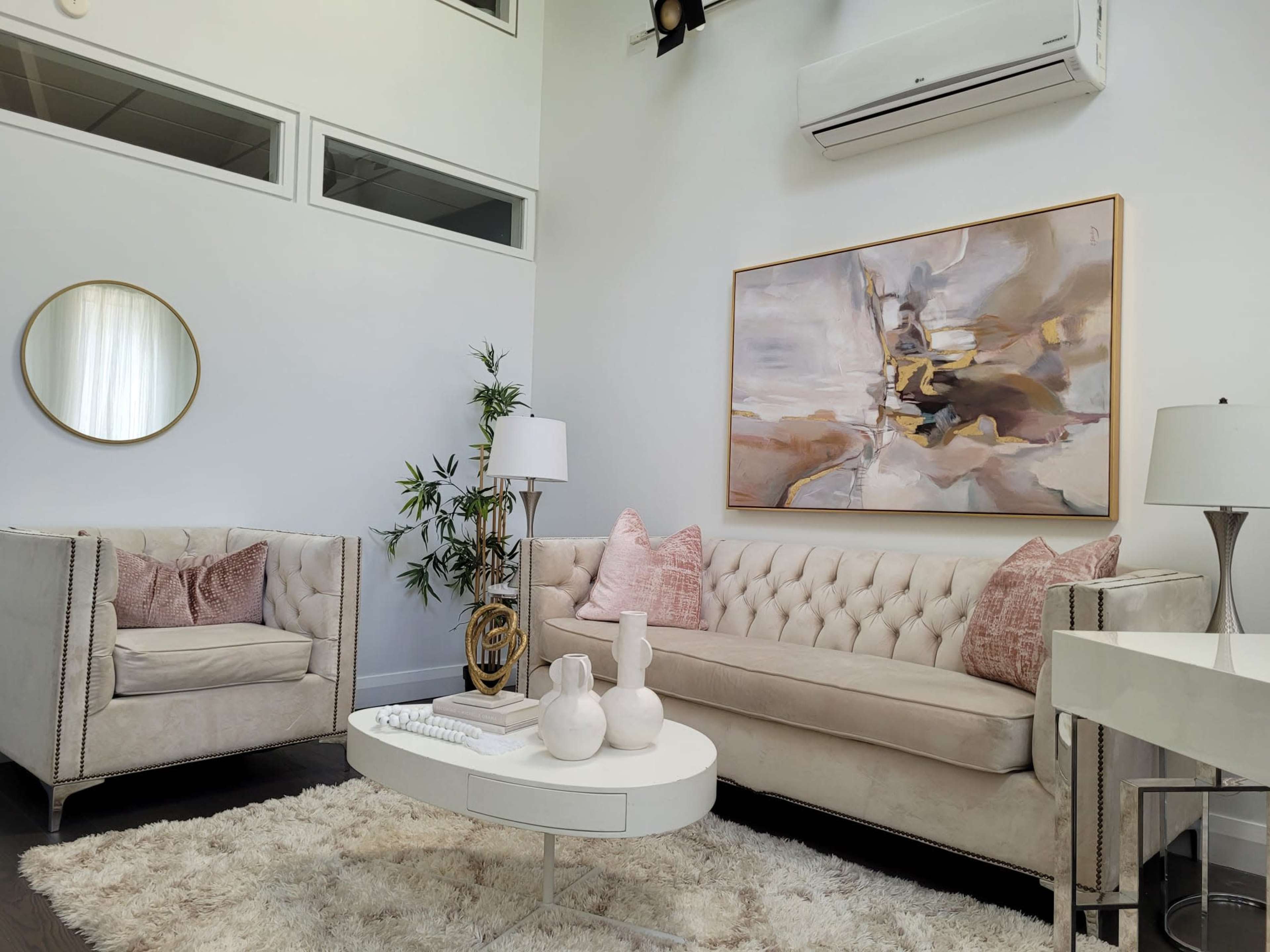 A bright living room with a beige tufted sofa, two armchairs, a round coffee table, and a large abstract painting on the wall.