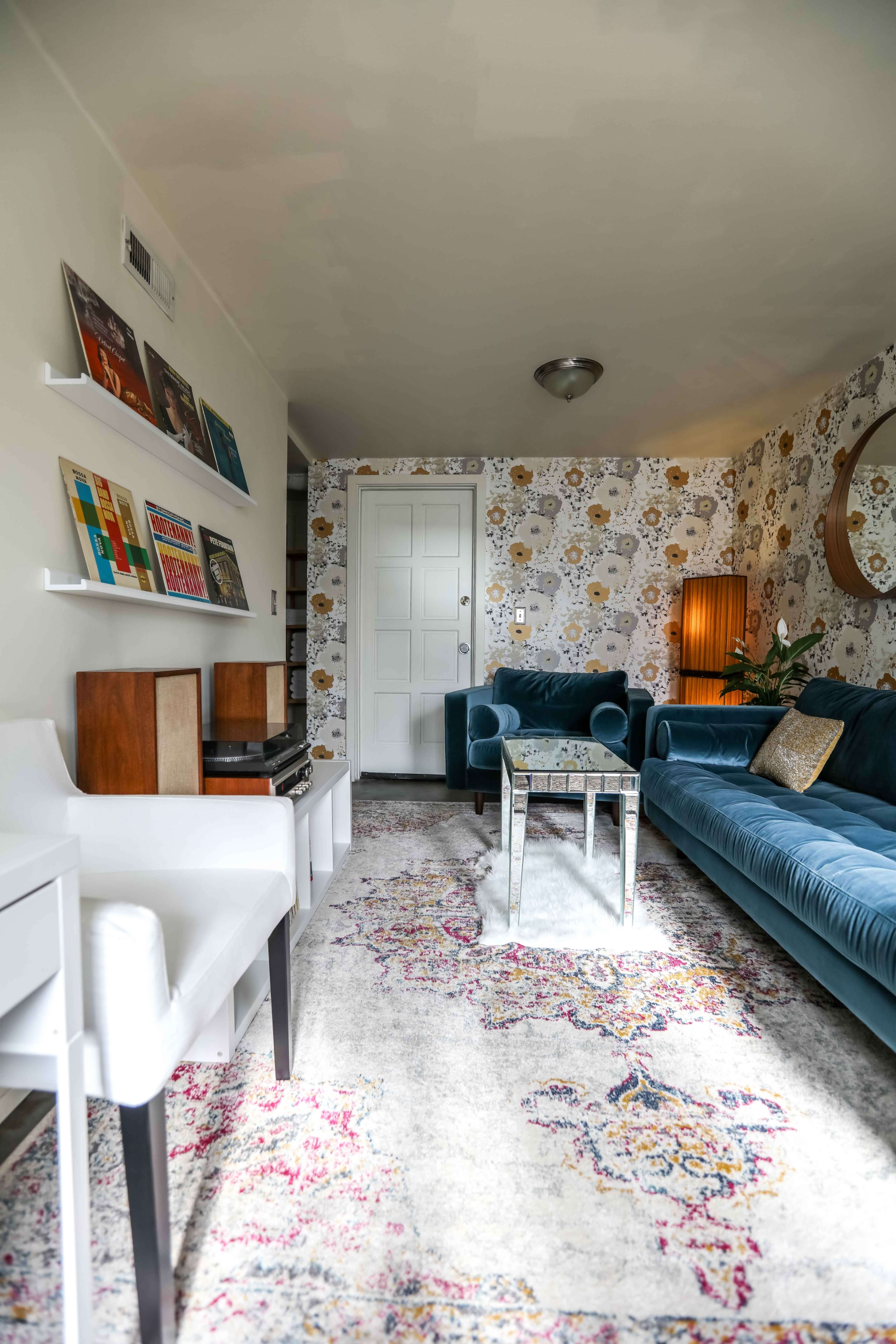 A living room features a blue sofa, a glass coffee table, a white desk chair, and floral wallpaper, with vinyl records displayed on a shelf.