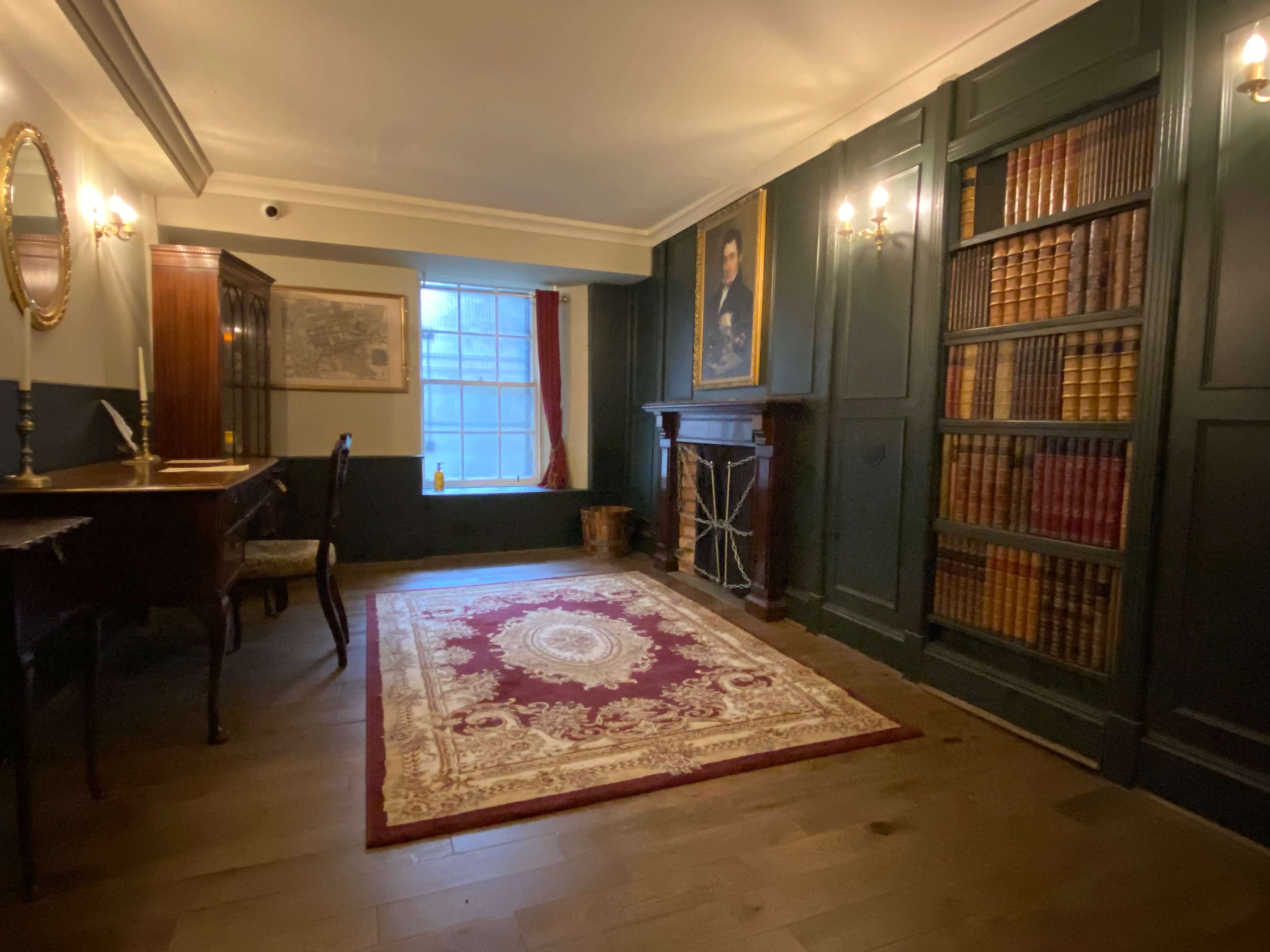 The image shows a dimly lit room featuring a wooden desk, a large bookshelf filled with books, a fireplace, and a decorative rug on the floor.