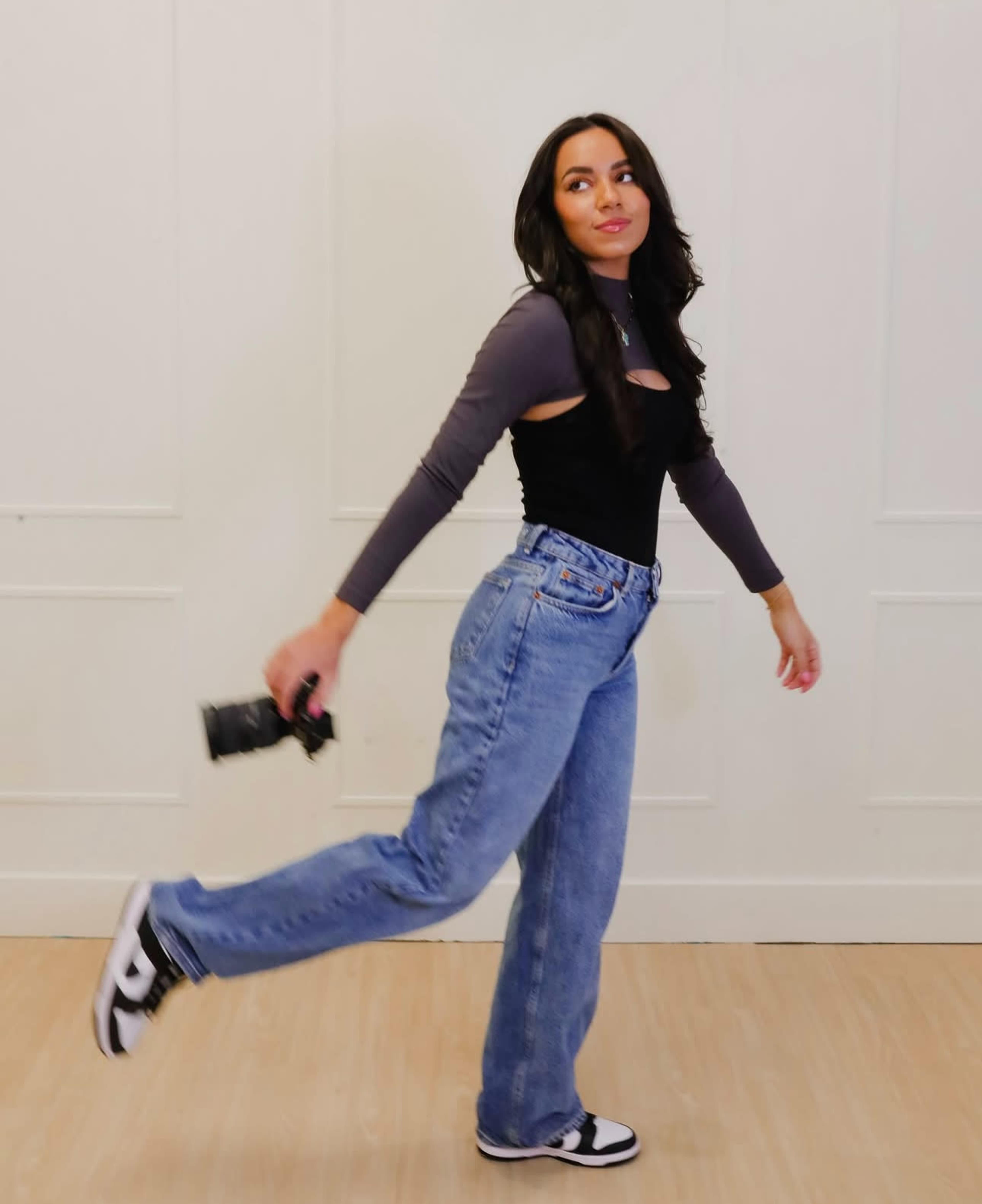 A person is walking across a room while holding a camera, wearing a black and gray outfit paired with loose blue jeans and sneakers.