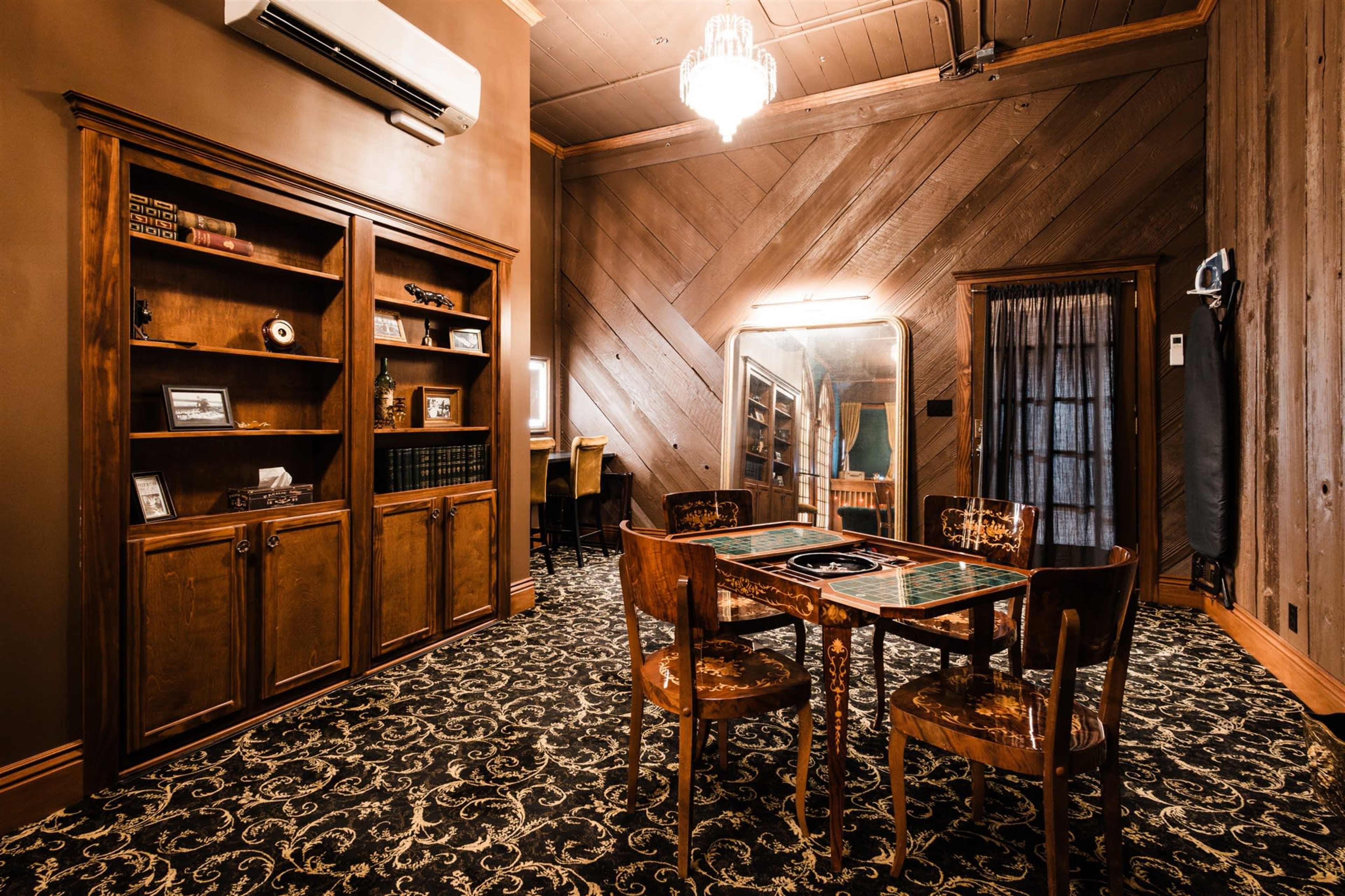 The image depicts a well-furnished room with wooden walls, a table in the center surrounded by chairs, a mirror on one wall, and a bookshelf filled with various items.