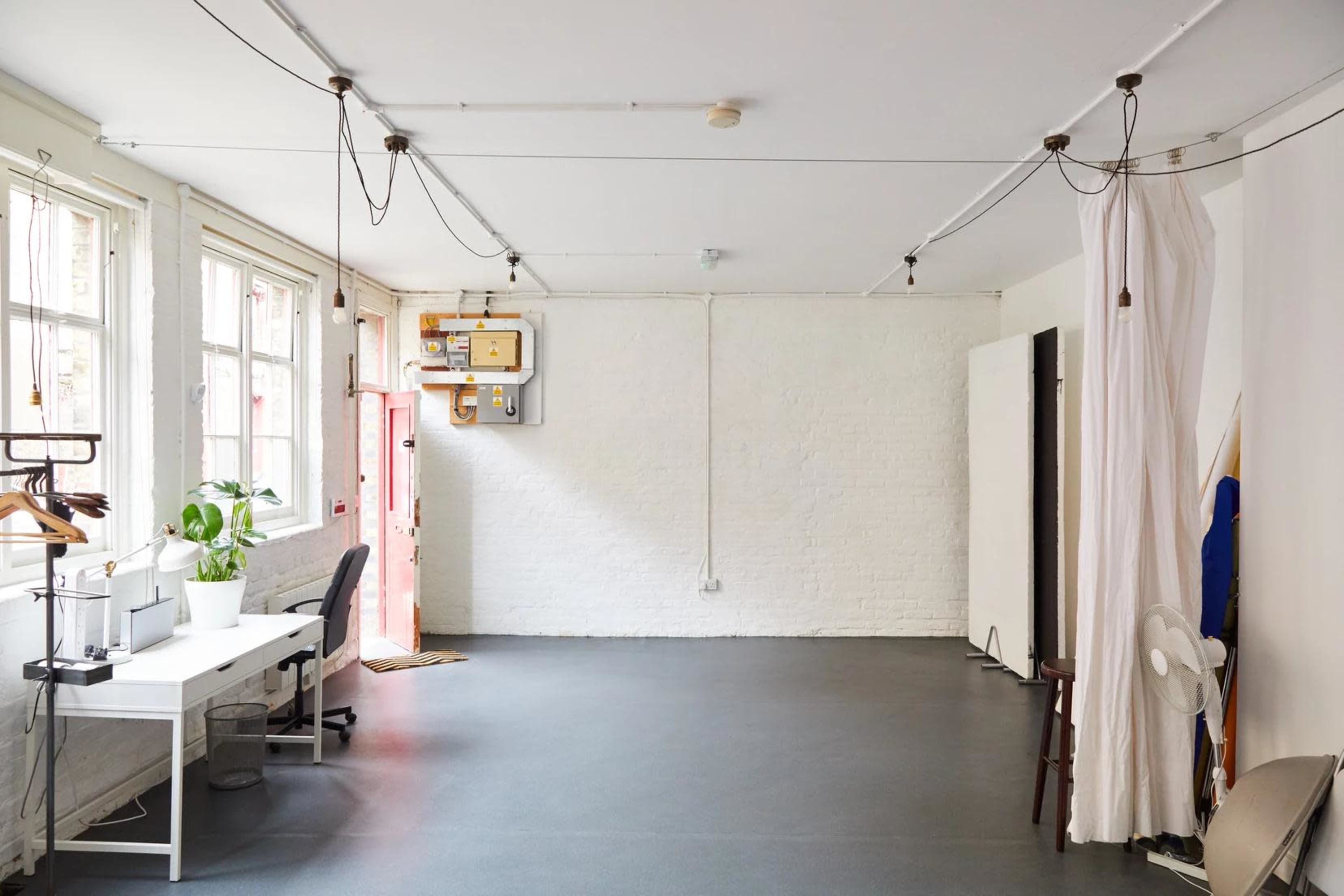 The image shows a spacious, empty room with large windows, a desk with two chairs, and a few furnishings including a plant and a curtain.