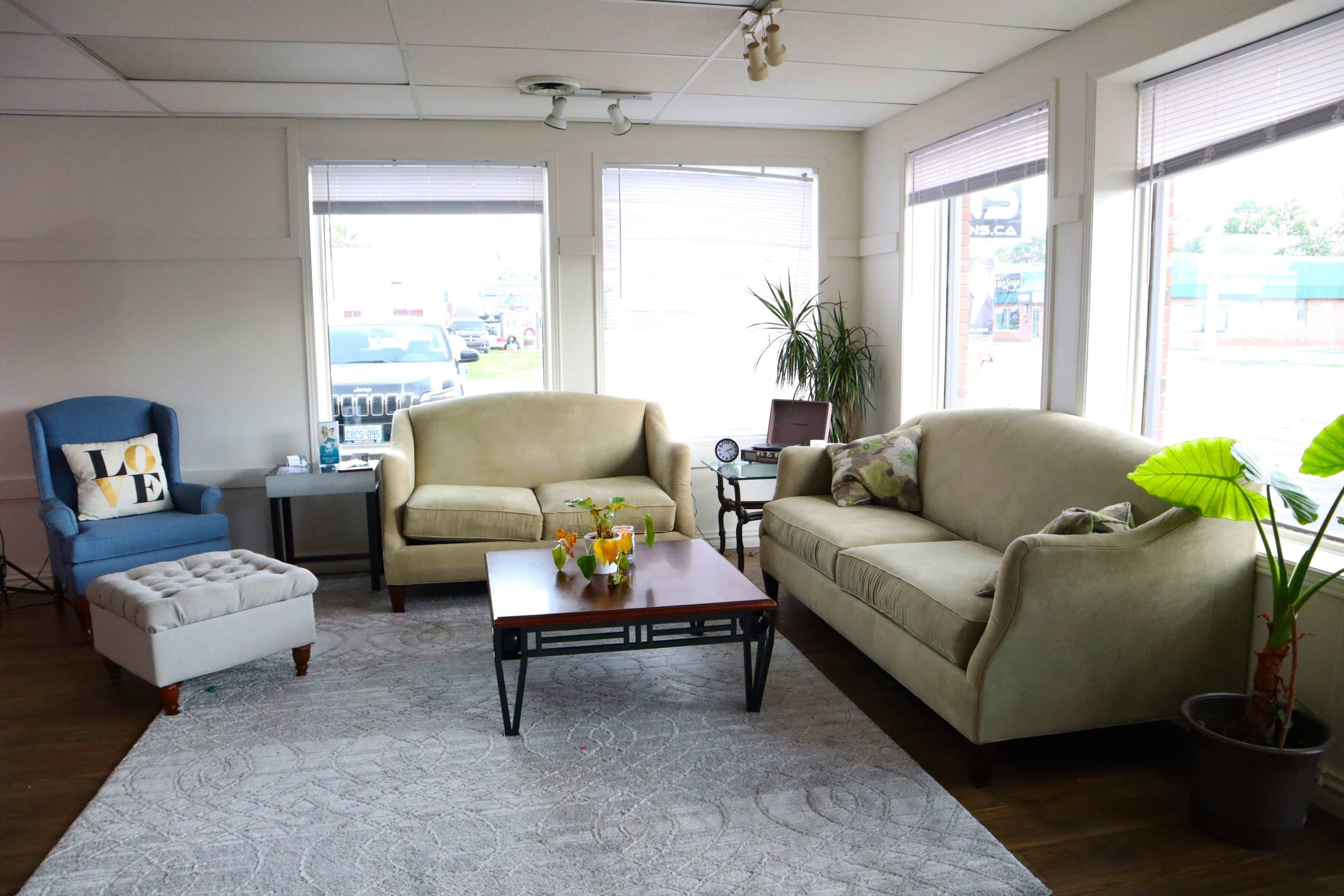 A well-lit living room features two sofas, a coffee table, and a small armchair, with large windows allowing natural light to enter.