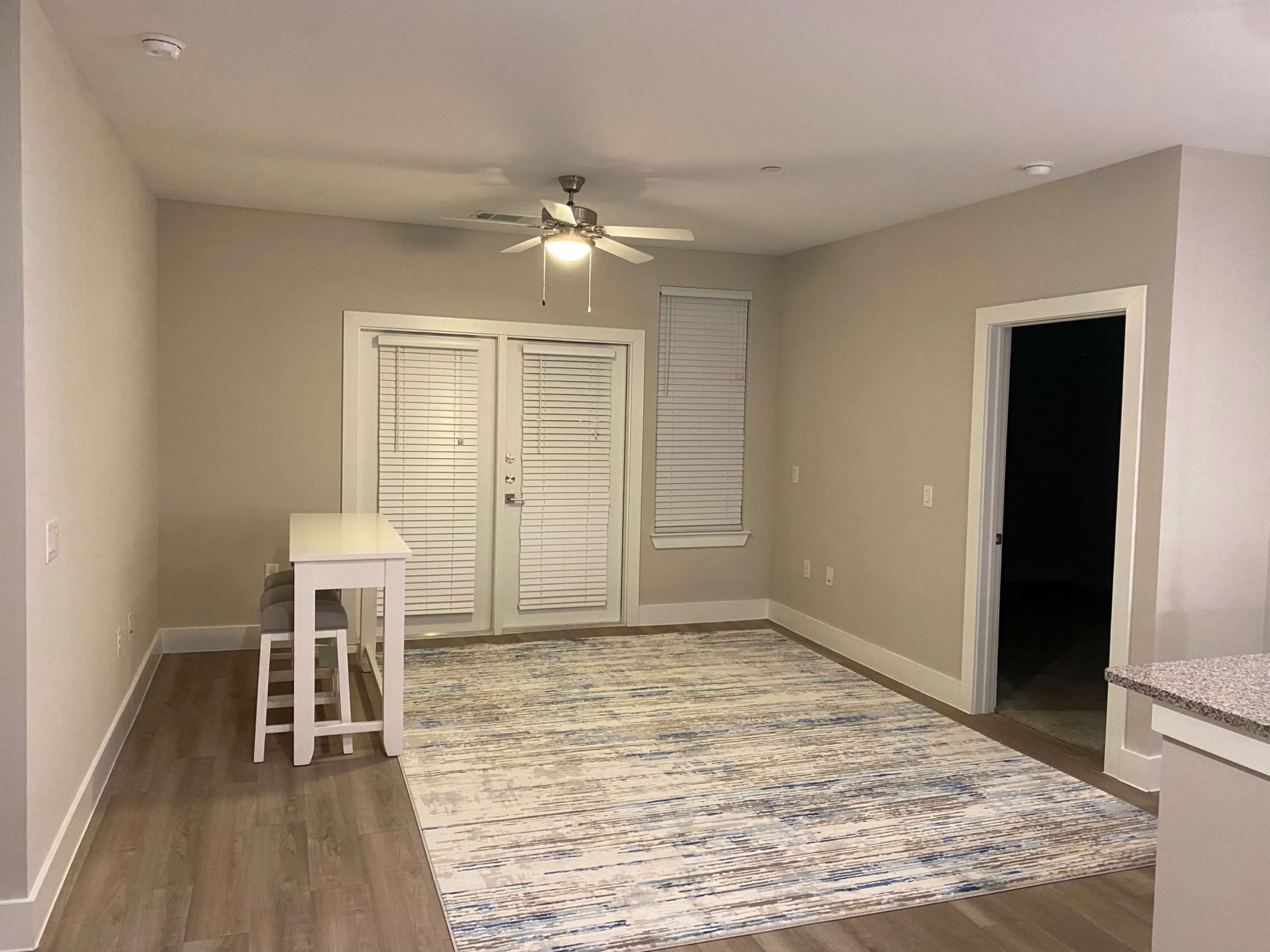 A spacious, well-lit room features a table with stools, a patterned rug, and large windows with blinds.