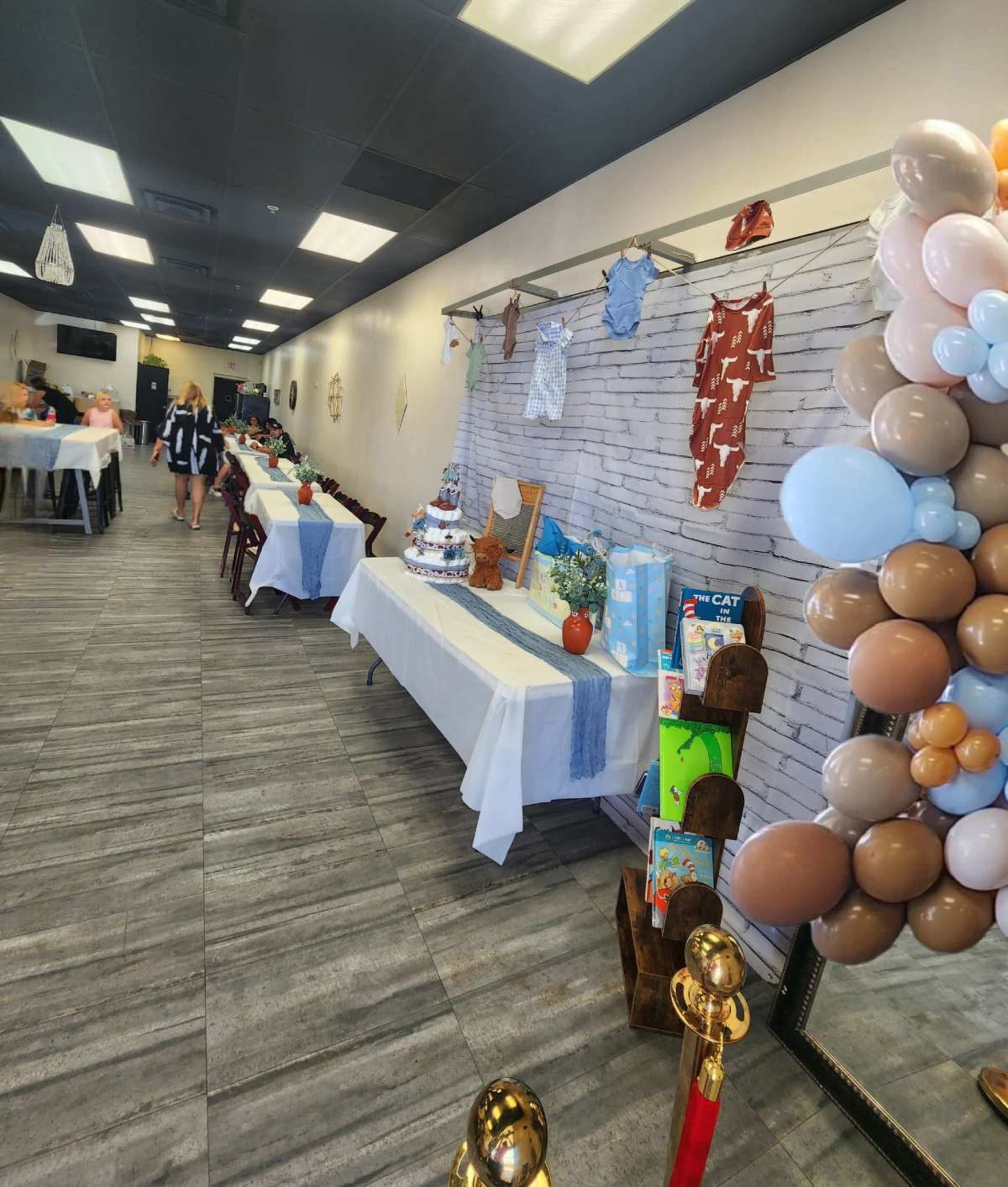 The image shows a decorated indoor event space with tables set up, a cake display at the front, and festive balloons near a mirror.