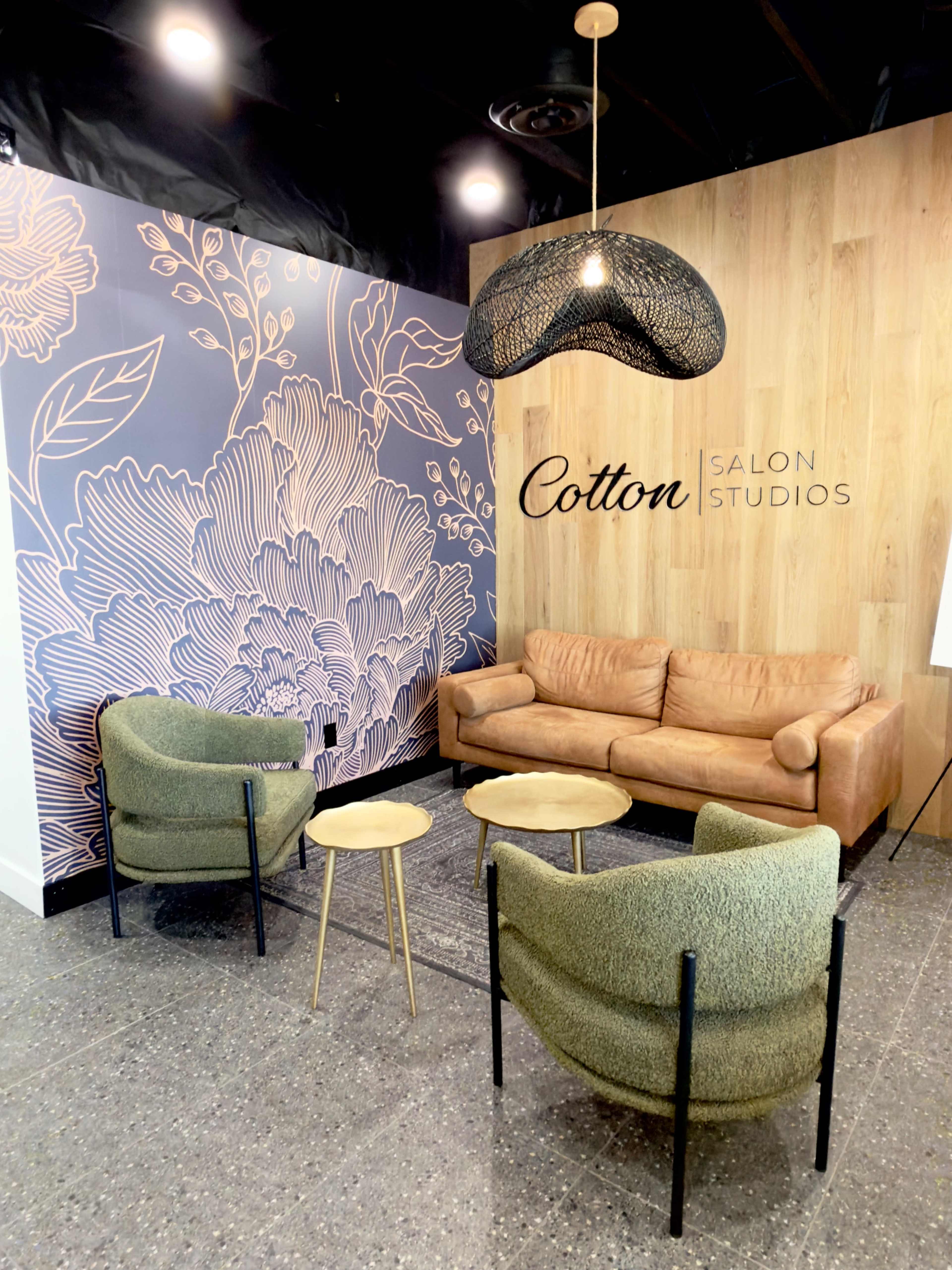 The image shows a waiting area in a salon featuring a brown leather couch, two green chairs, and a decorative wall with floral designs.