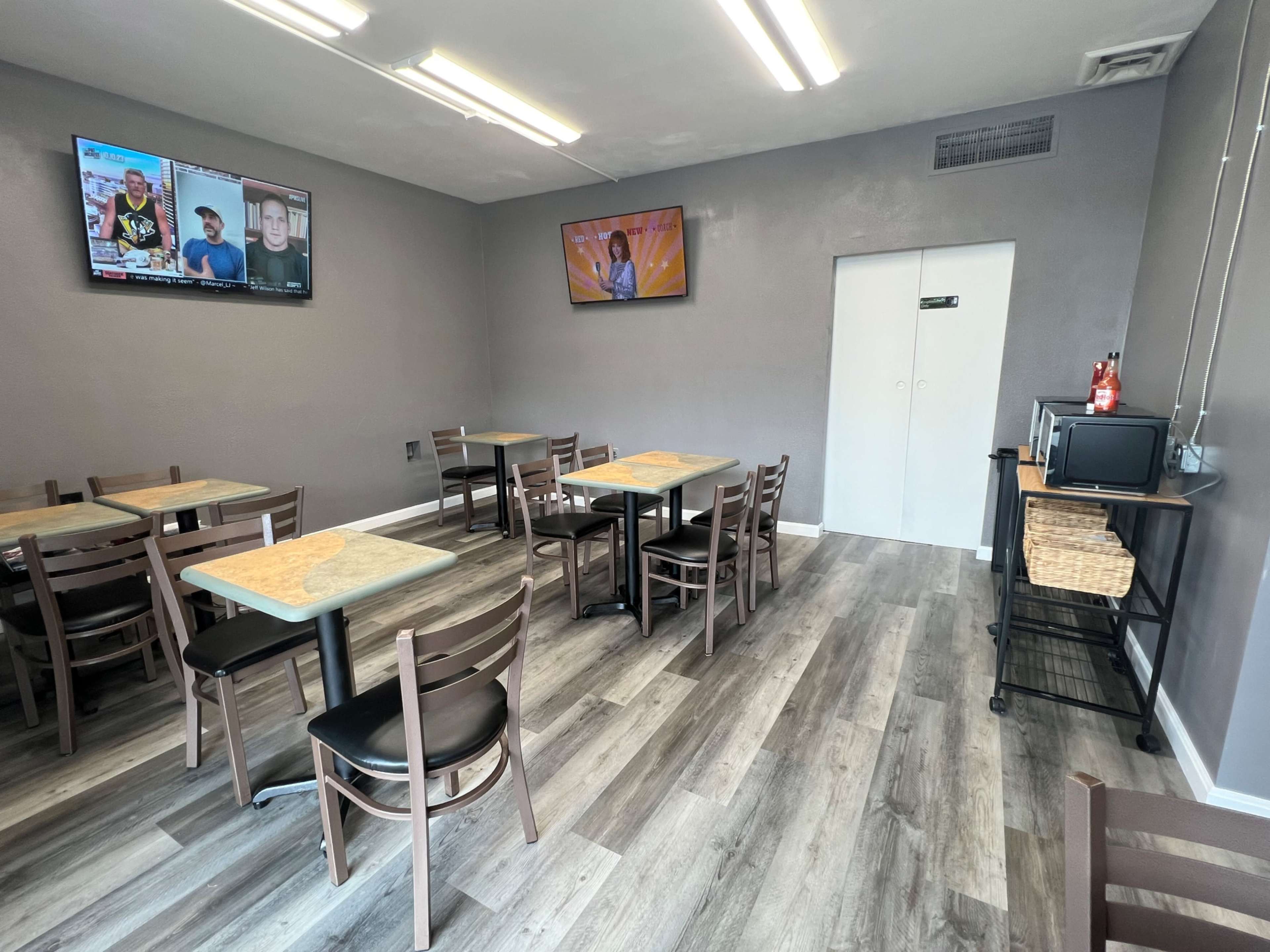 A simple dining area with several tables and chairs, two televisions mounted on the wall, and a microwave next to a storage cart.