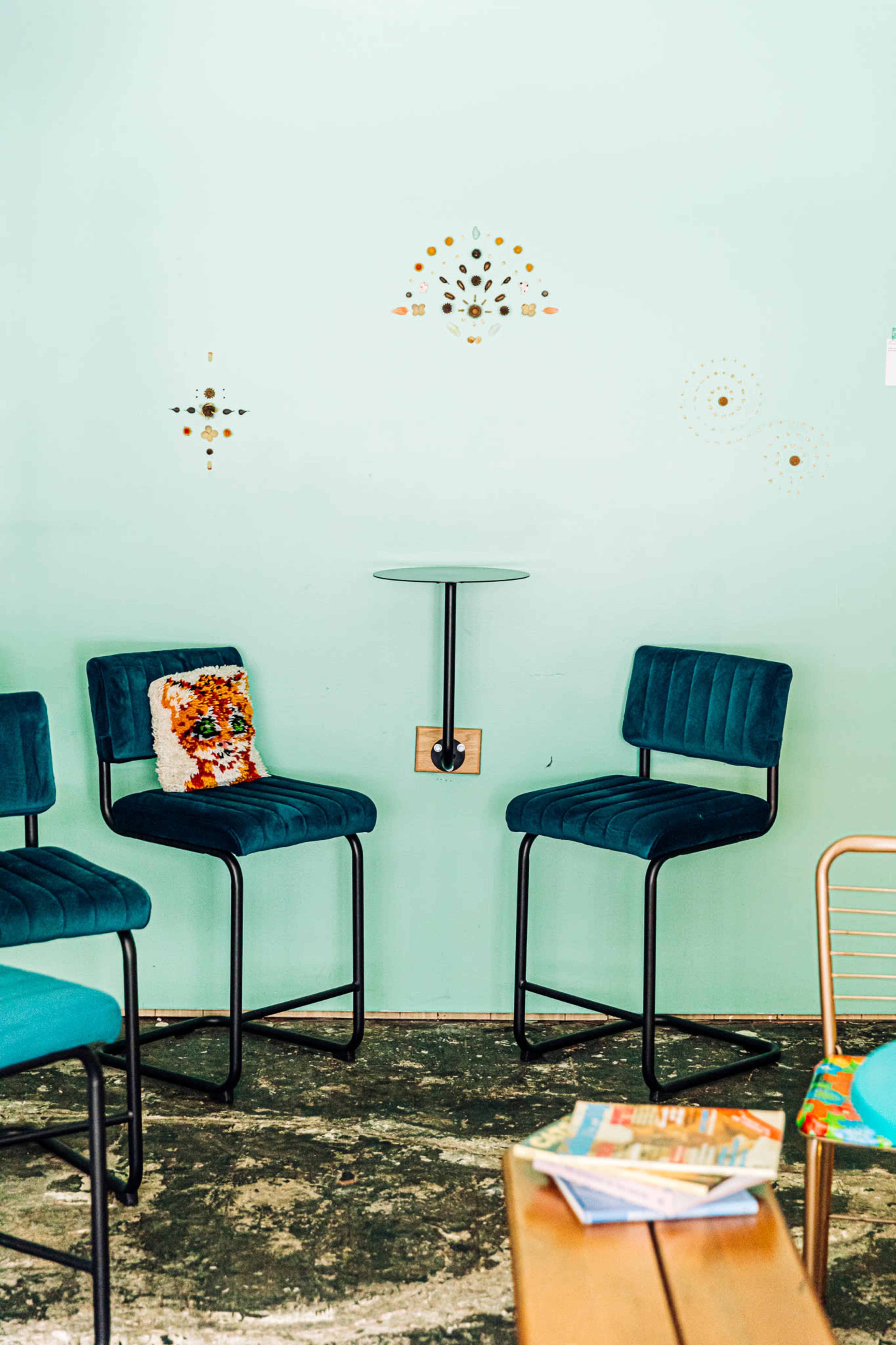 The image shows a corner of a room with two teal bar chairs, a small wall-mounted table, and decorative wall art in a light blue setting.