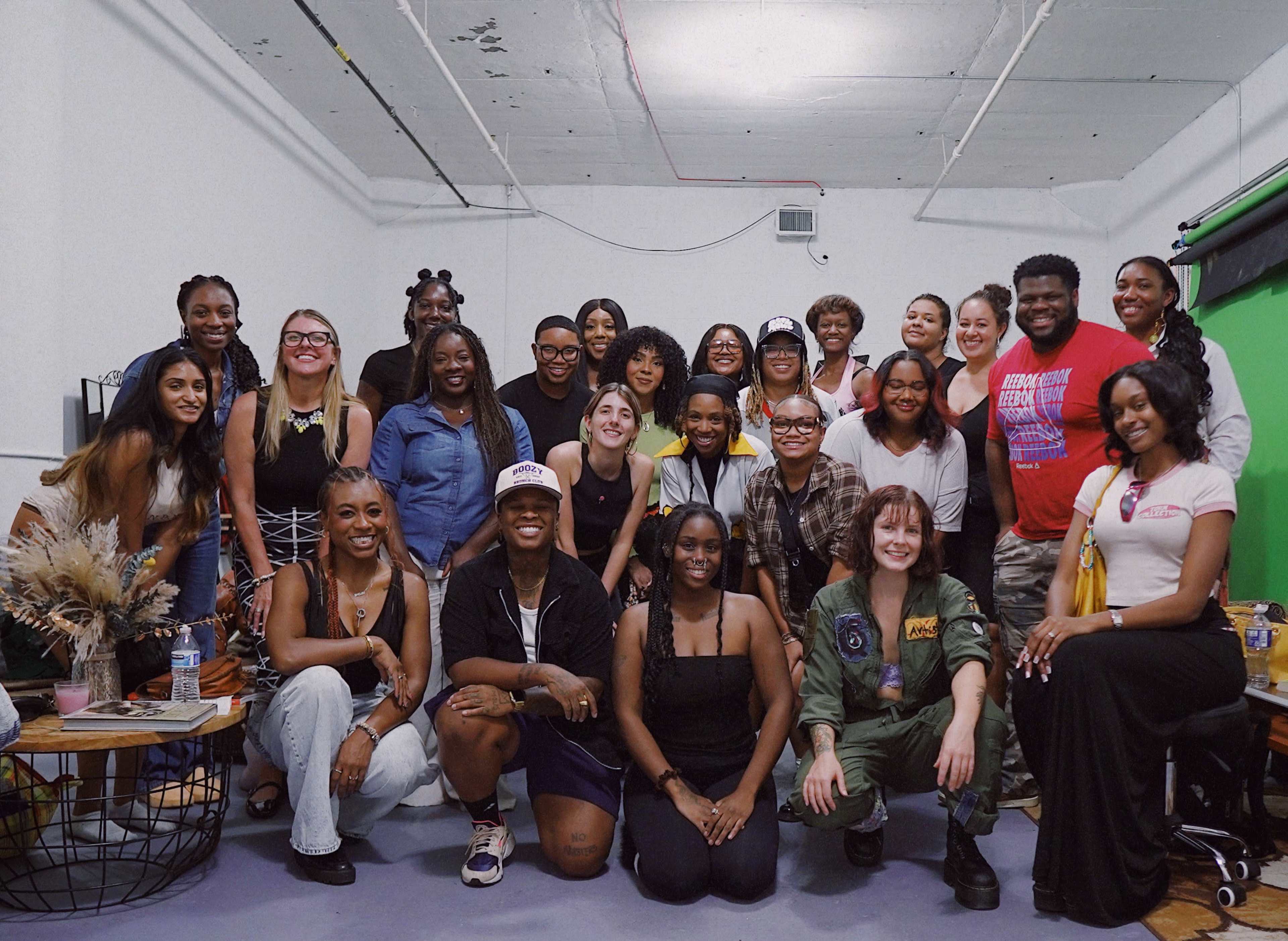 A diverse group of individuals poses together in a studio setting, with a mix of clothing styles and smiles.