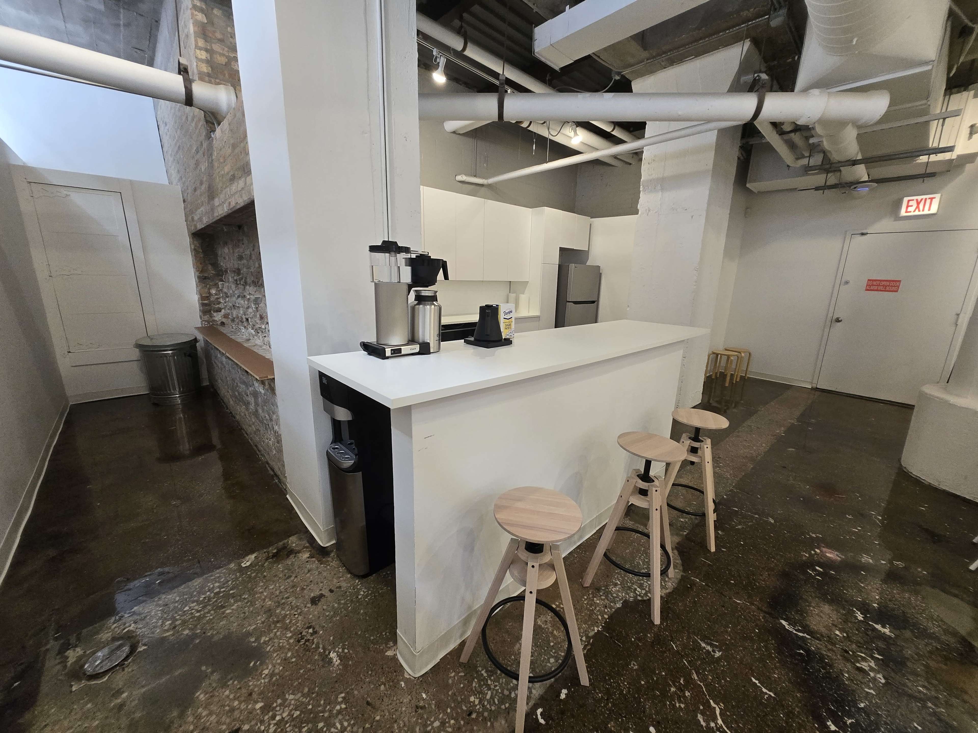 The image shows a modern kitchen area with a white countertop, three wooden stools, and stainless steel appliances in an industrial-style space.