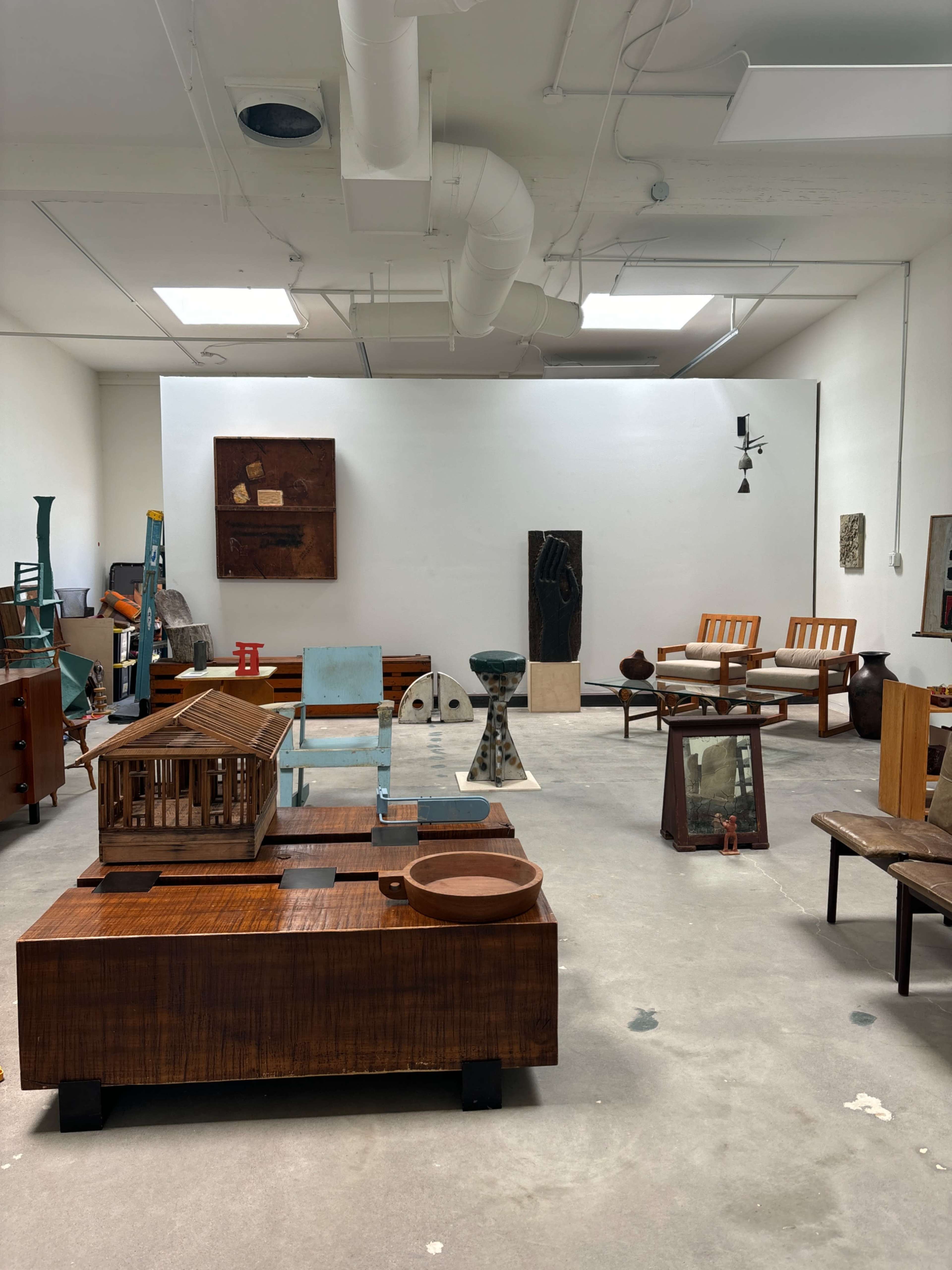 An art gallery or studio featuring a variety of wooden sculptures and furniture pieces arranged on a concrete floor.