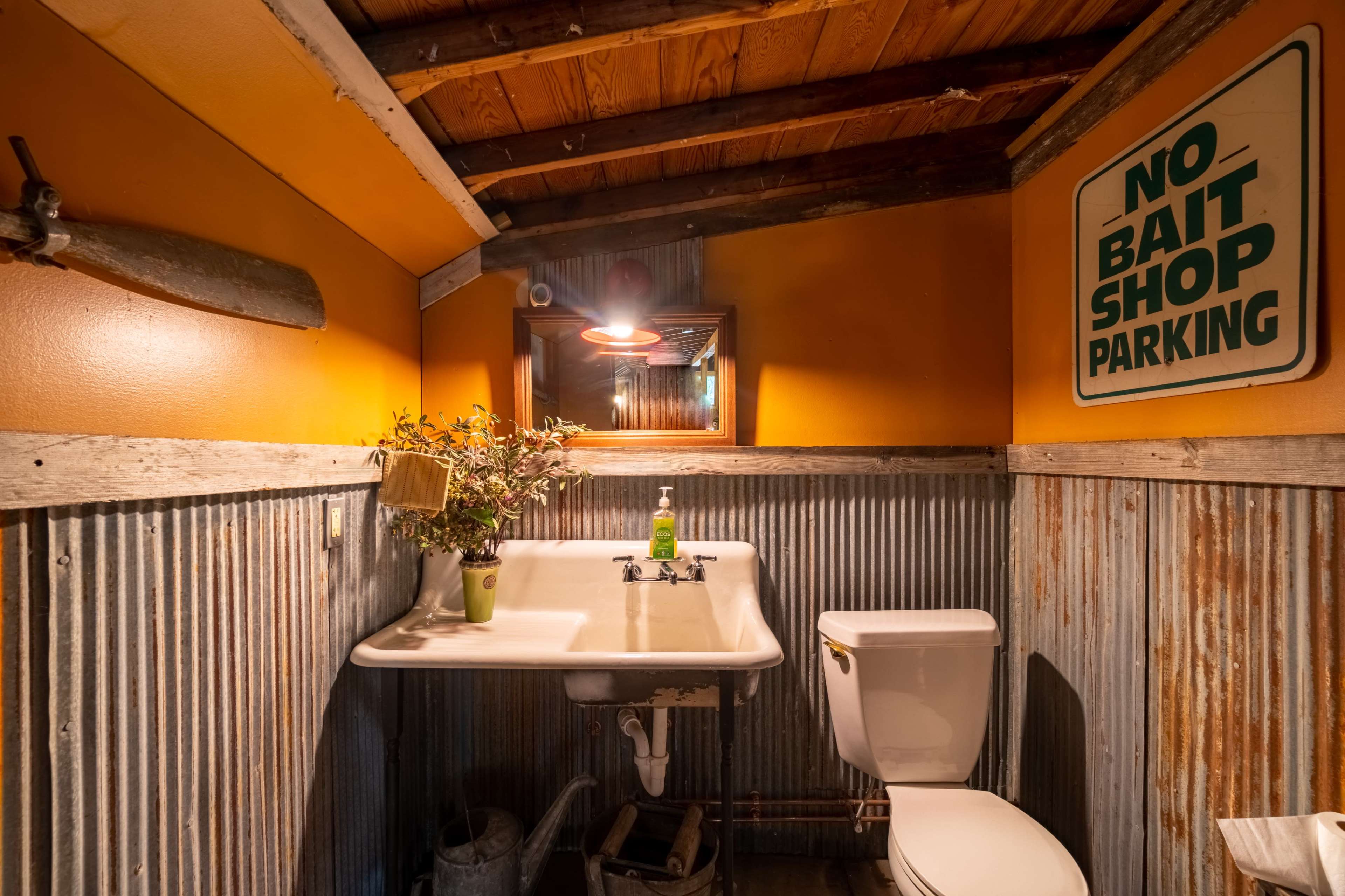 The image shows a rustic bathroom with a sink, toilet, and decorative elements such as a plants and a sign on the walls.