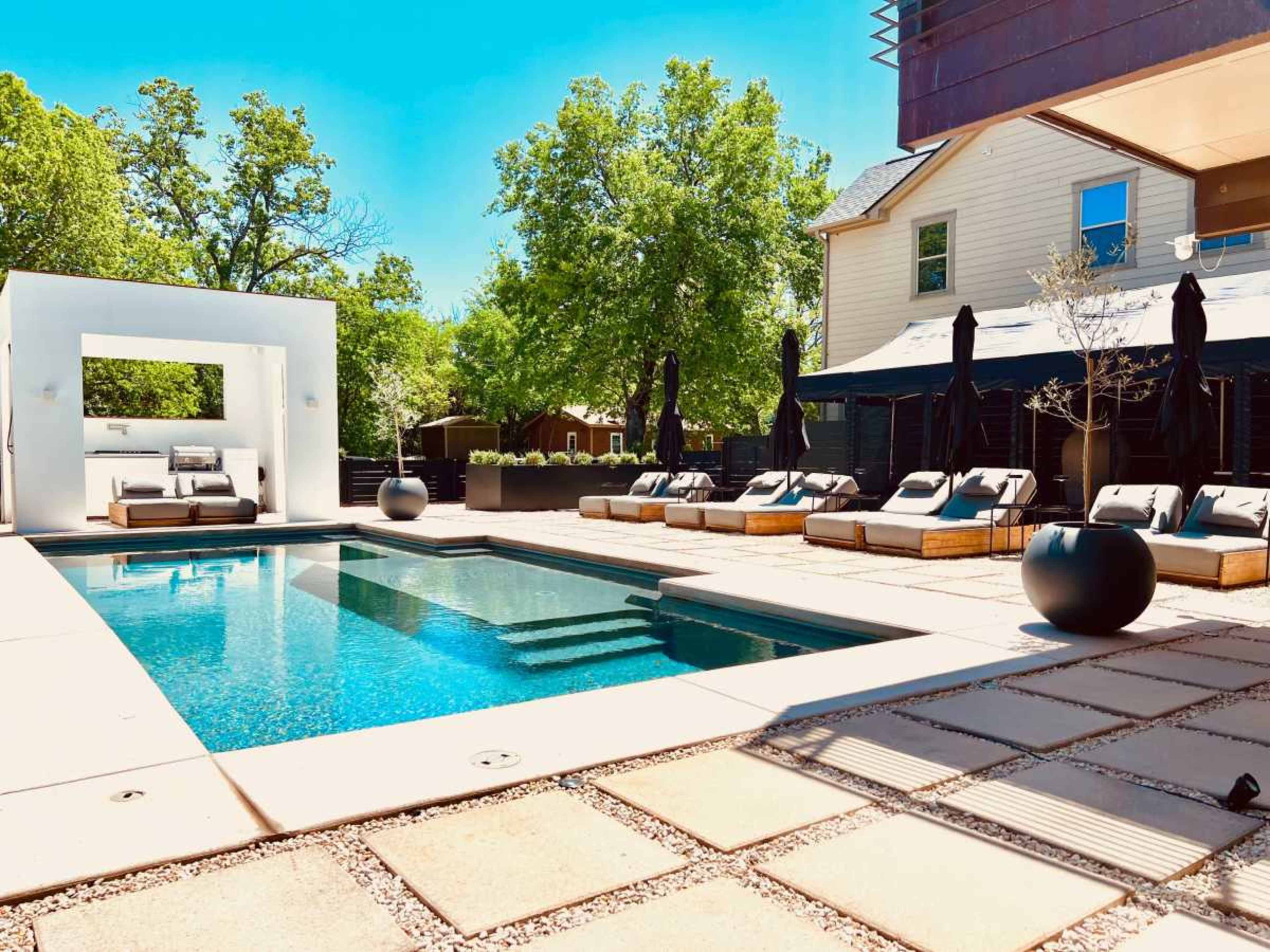 The image shows a modern outdoor pool area surrounded by lounge chairs, a sleek white structure, and lush green trees in the background.