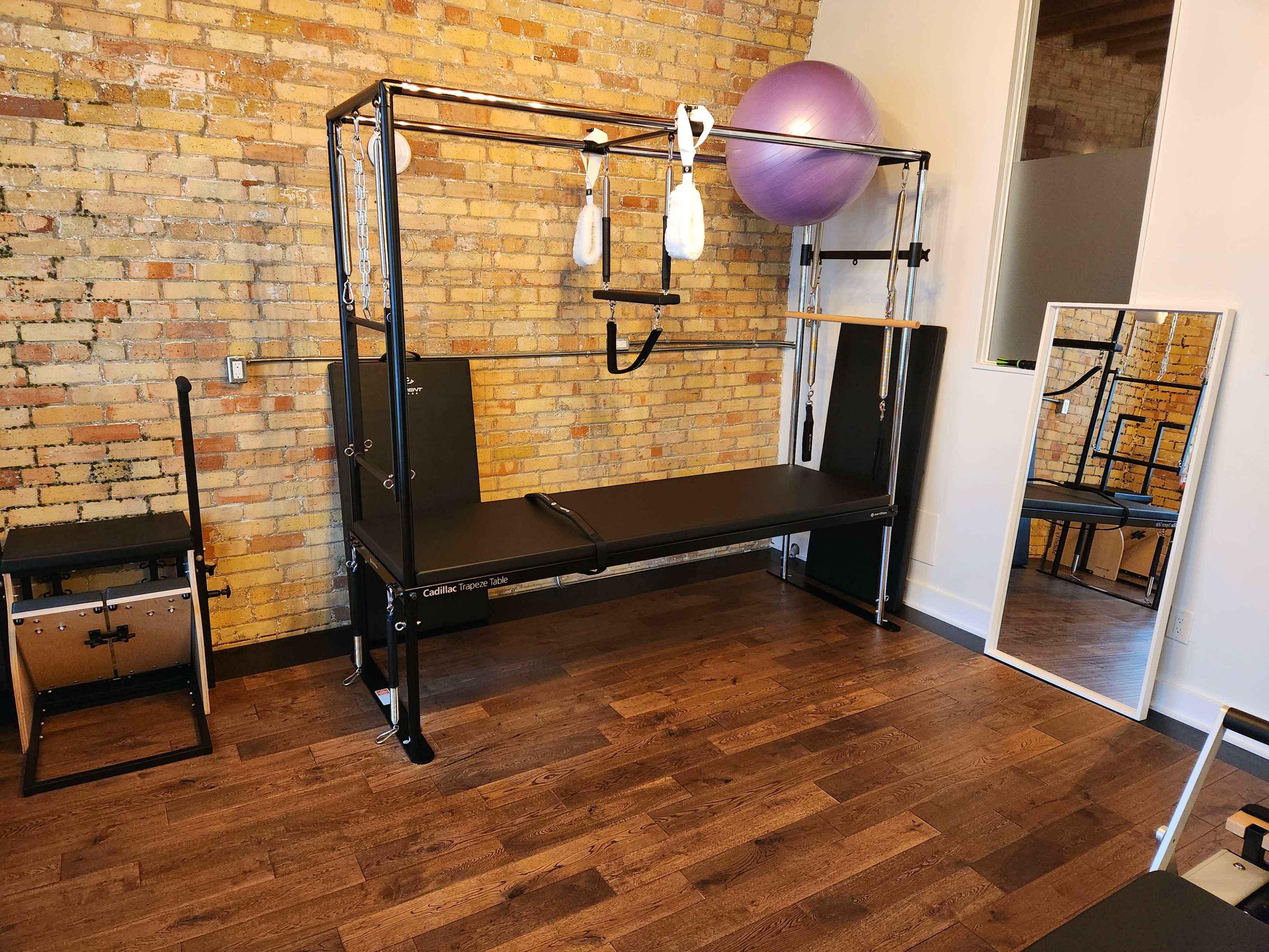 A Pilates reformer machine positioned against a brick wall, with a workout ball and mirror visible in the room.