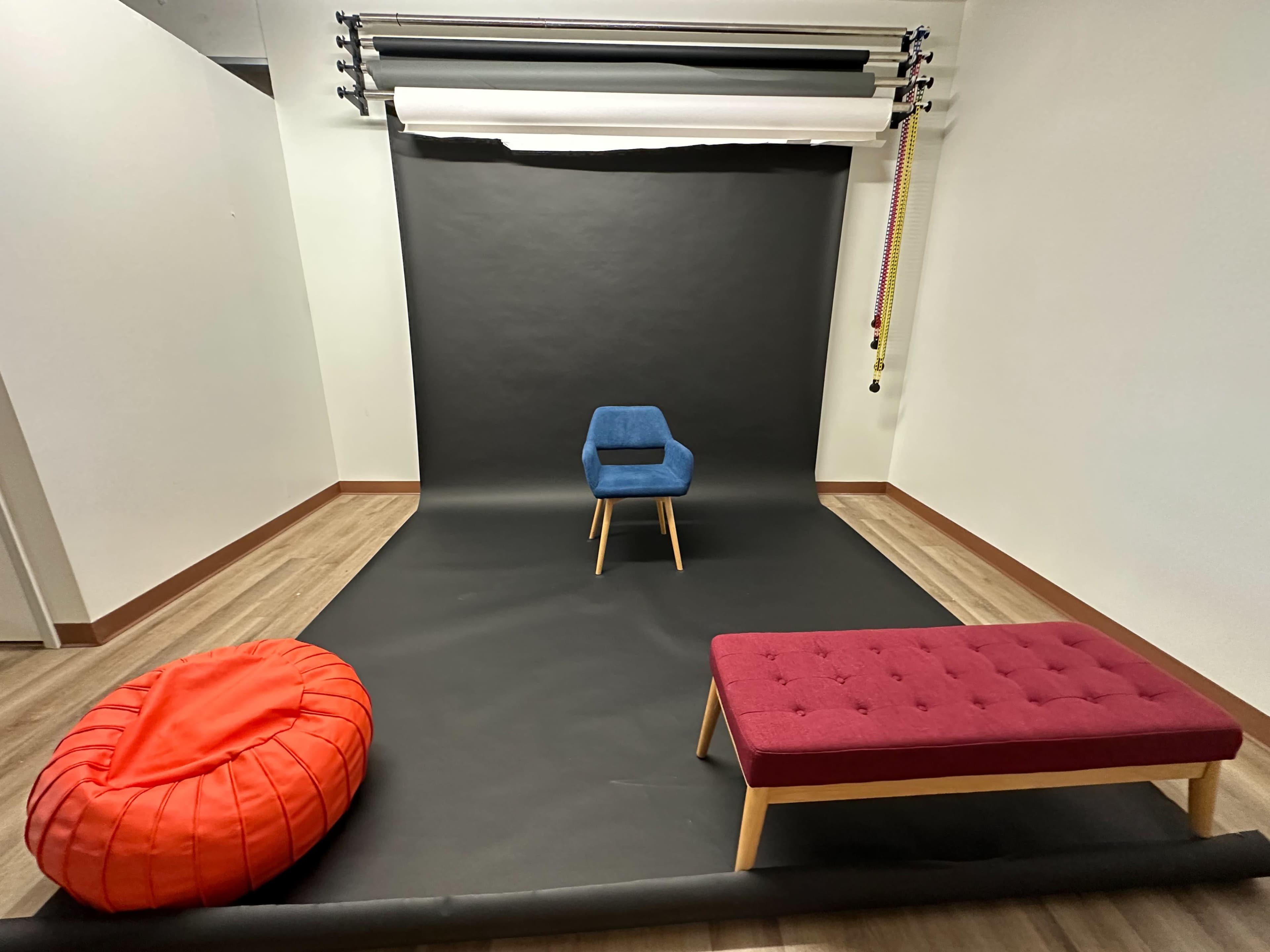 The image shows a minimalist studio setting with a blue chair, a red bean bag, and a purple bench against a dark backdrop on a wooden floor.