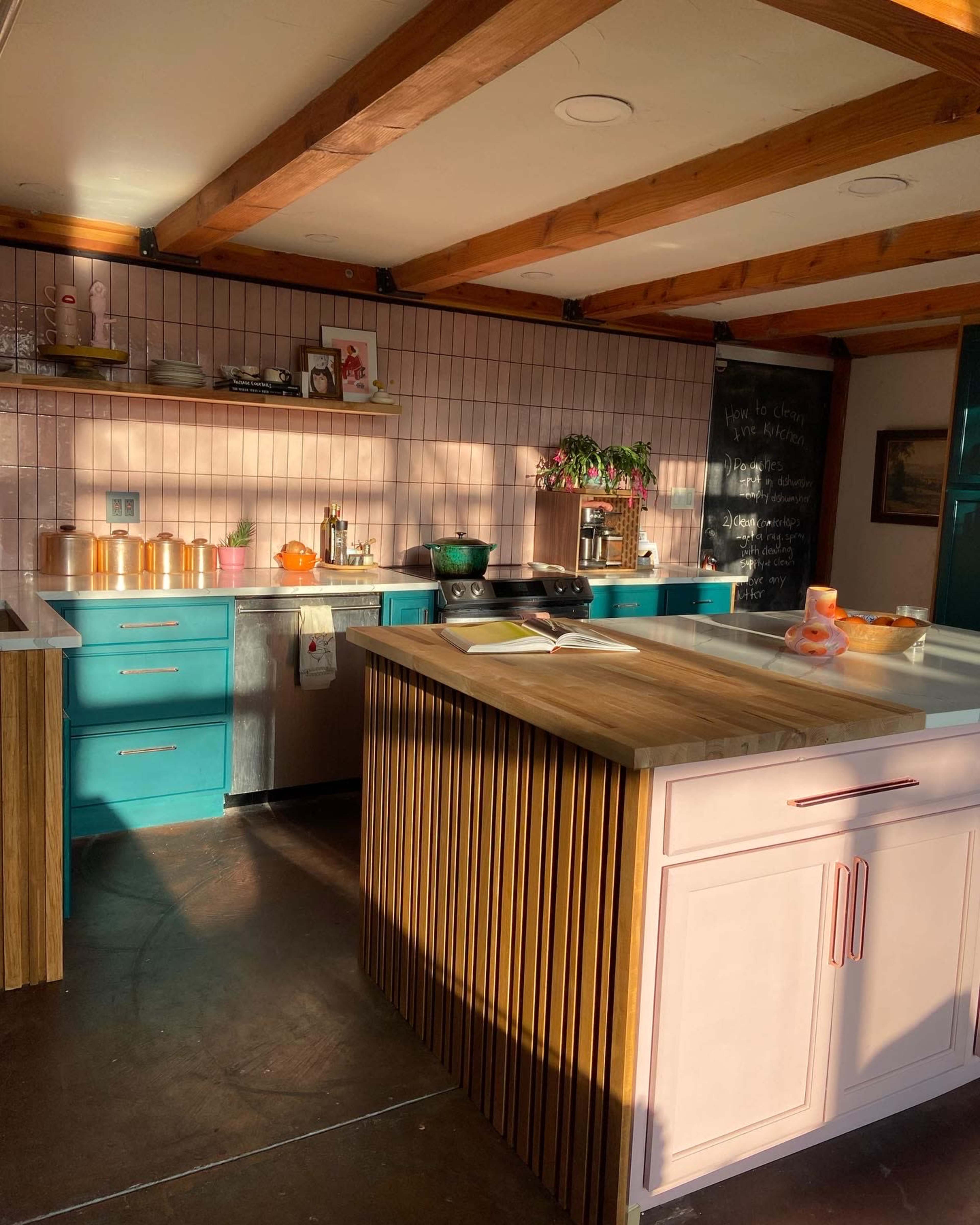 The kitchen features teal cabinets, a pink island with wooden accents, and ample sunlight illuminating the space.