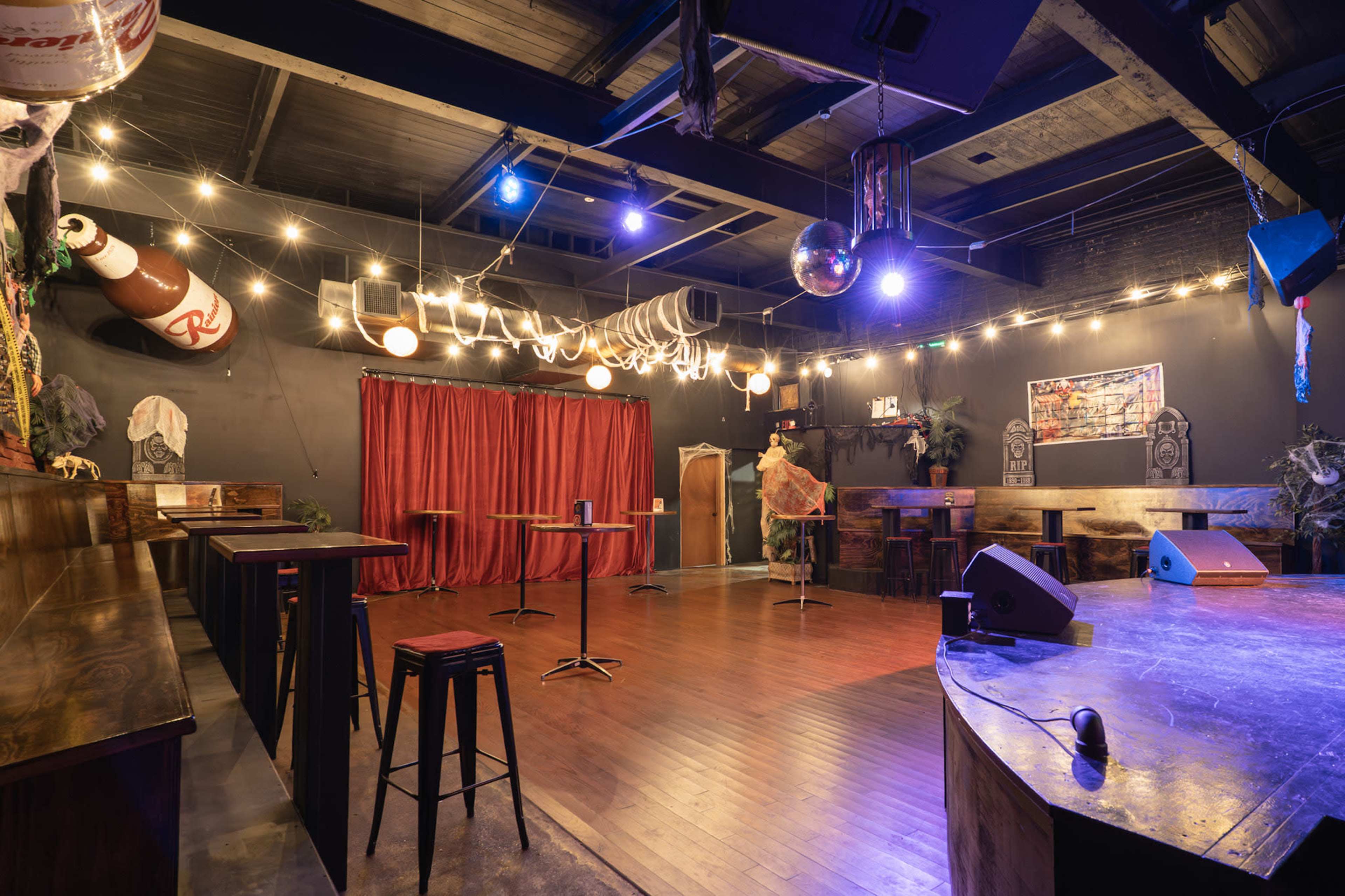 The image shows a dimly lit bar or performance space with a wooden floor, empty tables, and a red curtain backdrop, decorated with string lights and a disco ball.