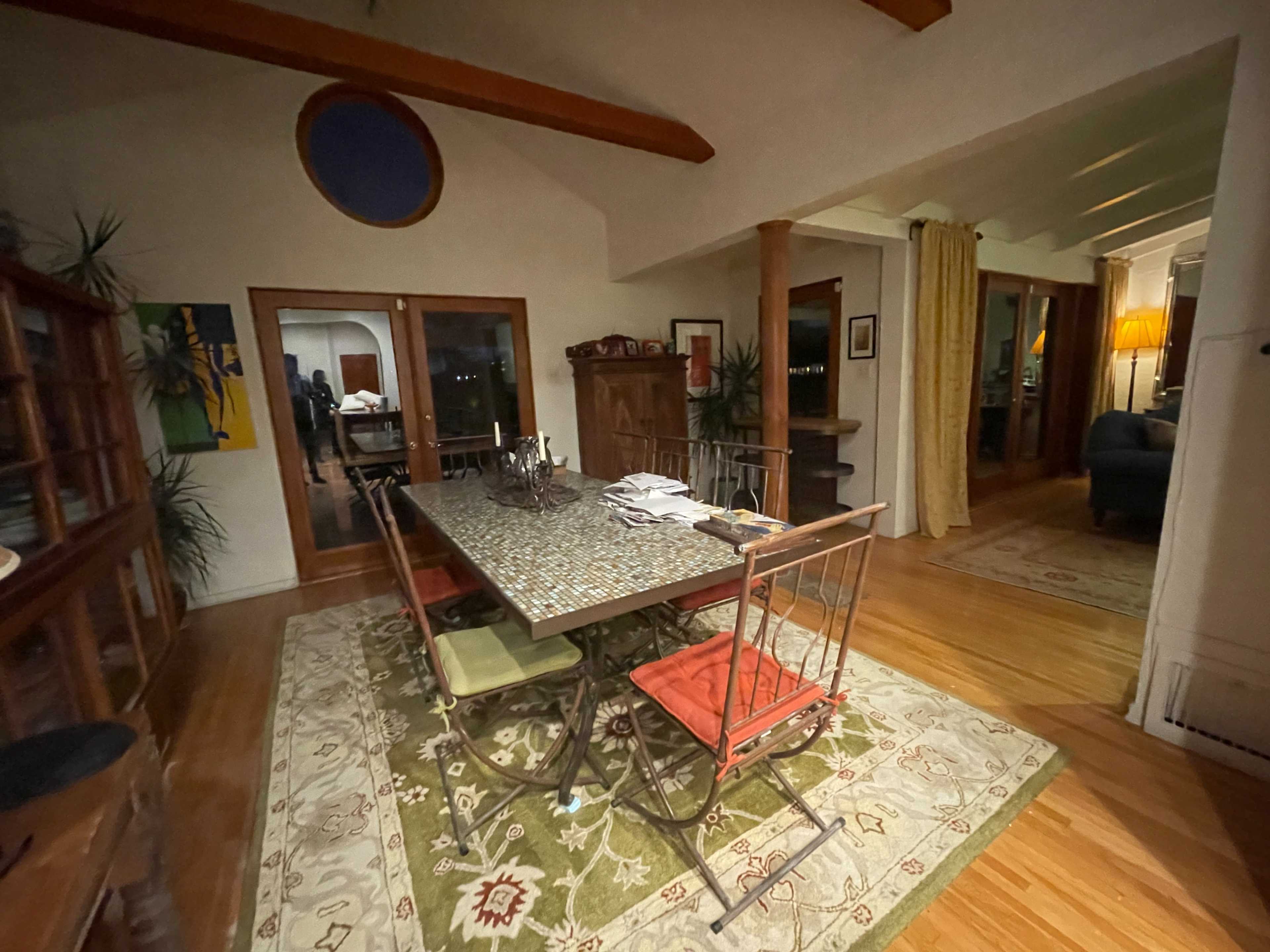 A dining area with a large table surrounded by chairs, set on a patterned rug, with windows and a warm lamp in the background.