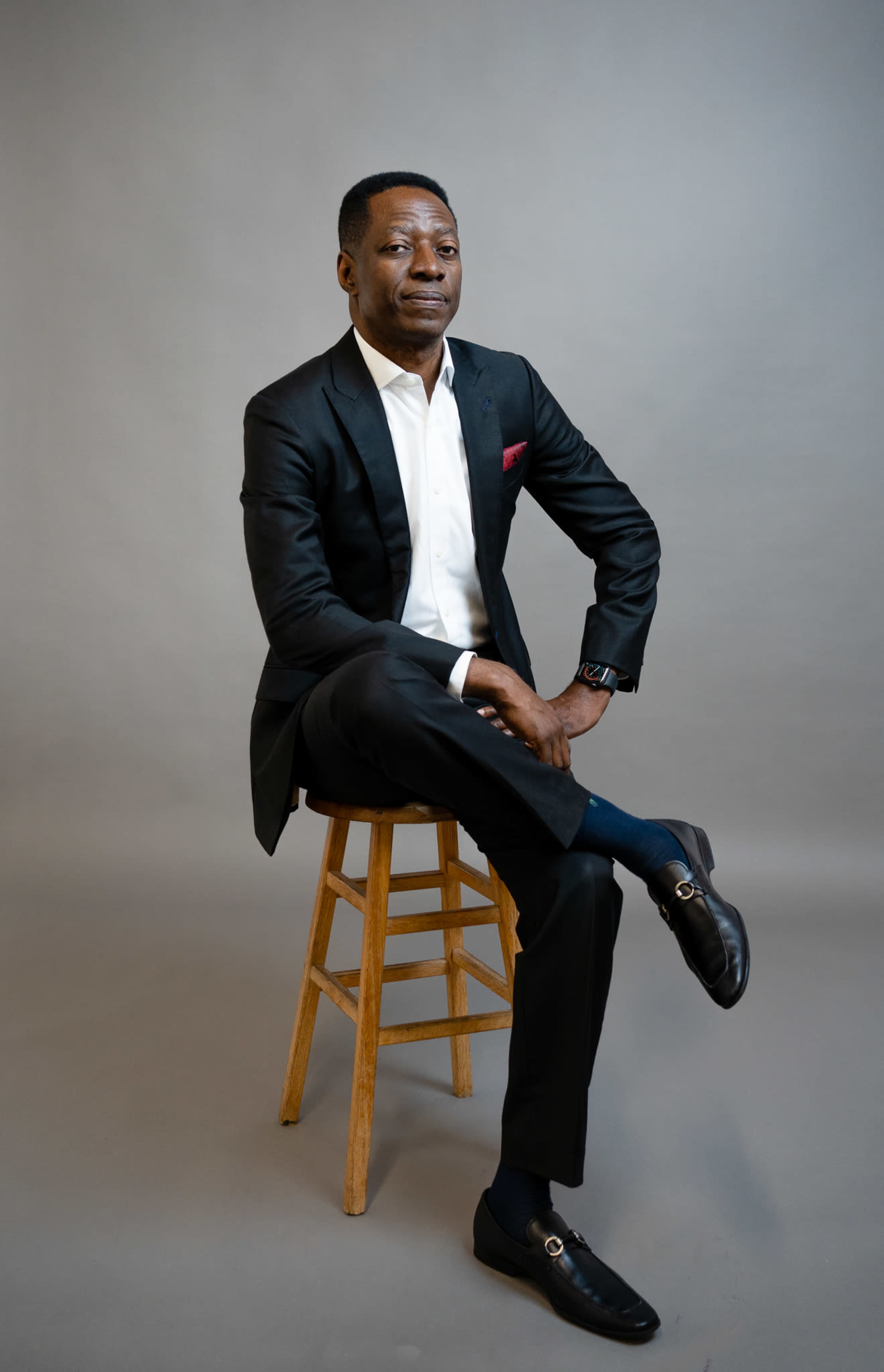 A man in a black suit and white shirt sits on a wooden stool with one leg crossed over the other.