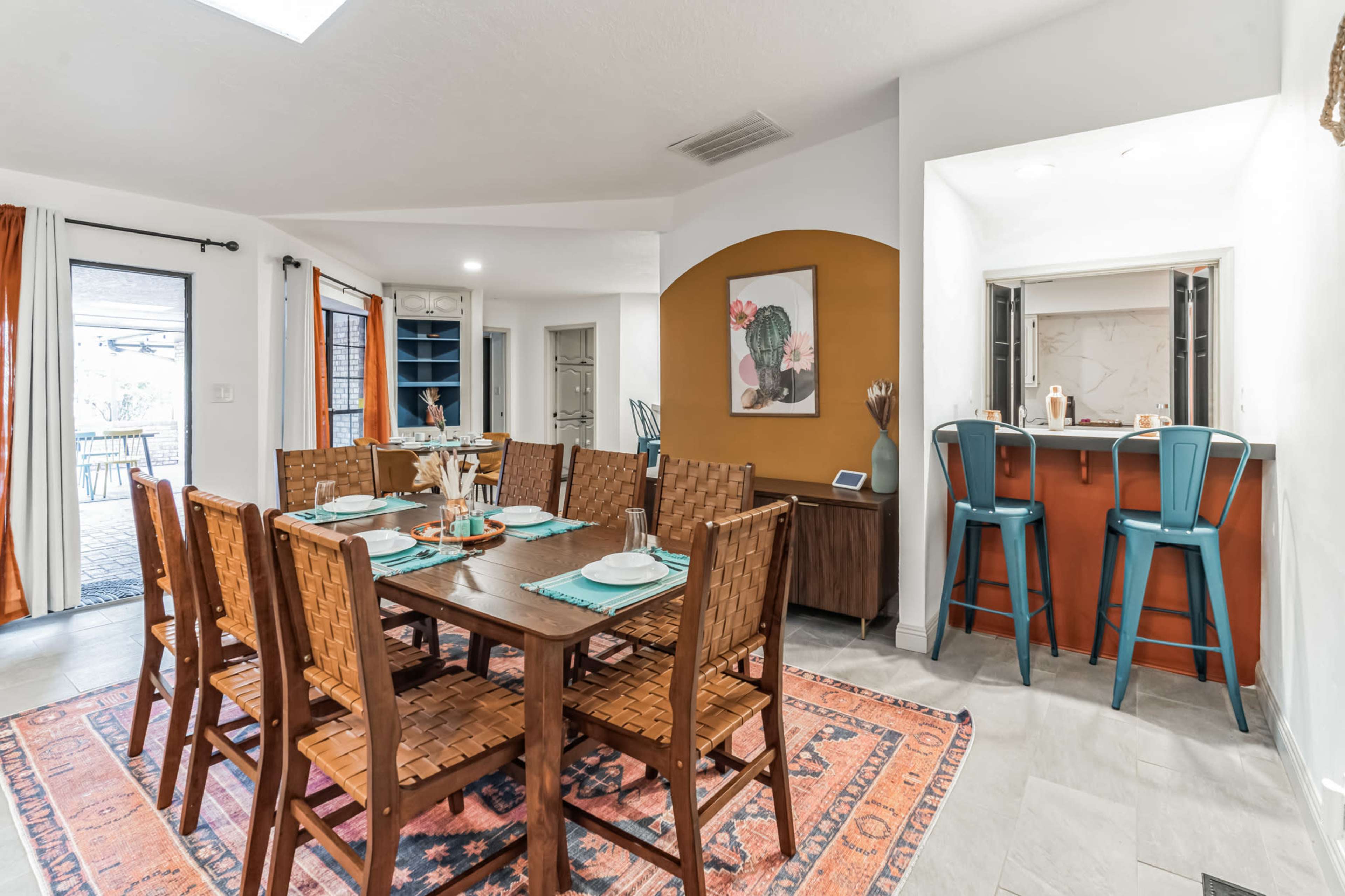 A spacious dining area features a large wooden table set for a meal, surrounded by woven chairs, with a kitchenette visible in the background.