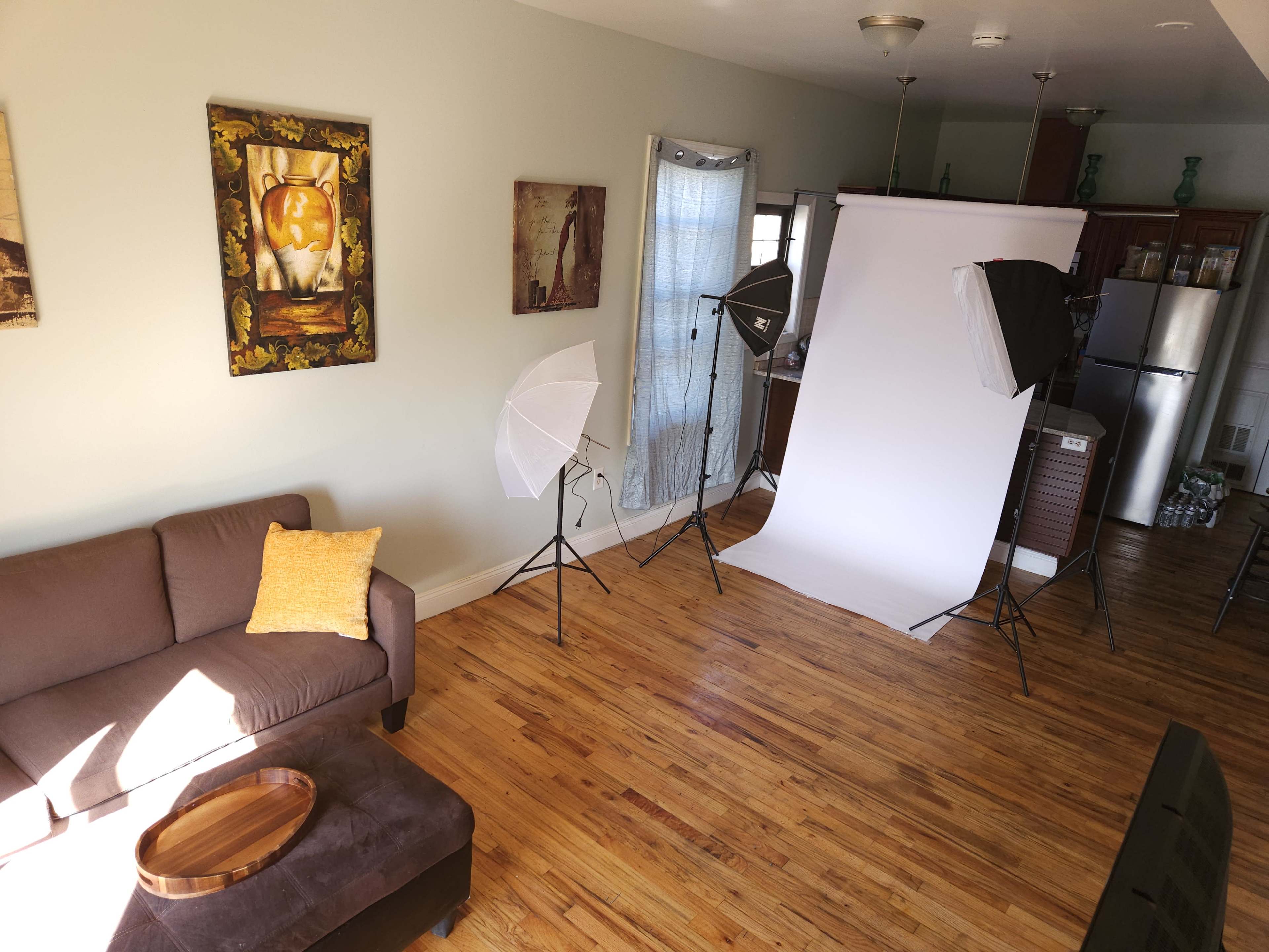 The image shows a bright living room with a brown couch, a wooden coffee table, and two softbox lights set up to illuminate a backdrop for photography.