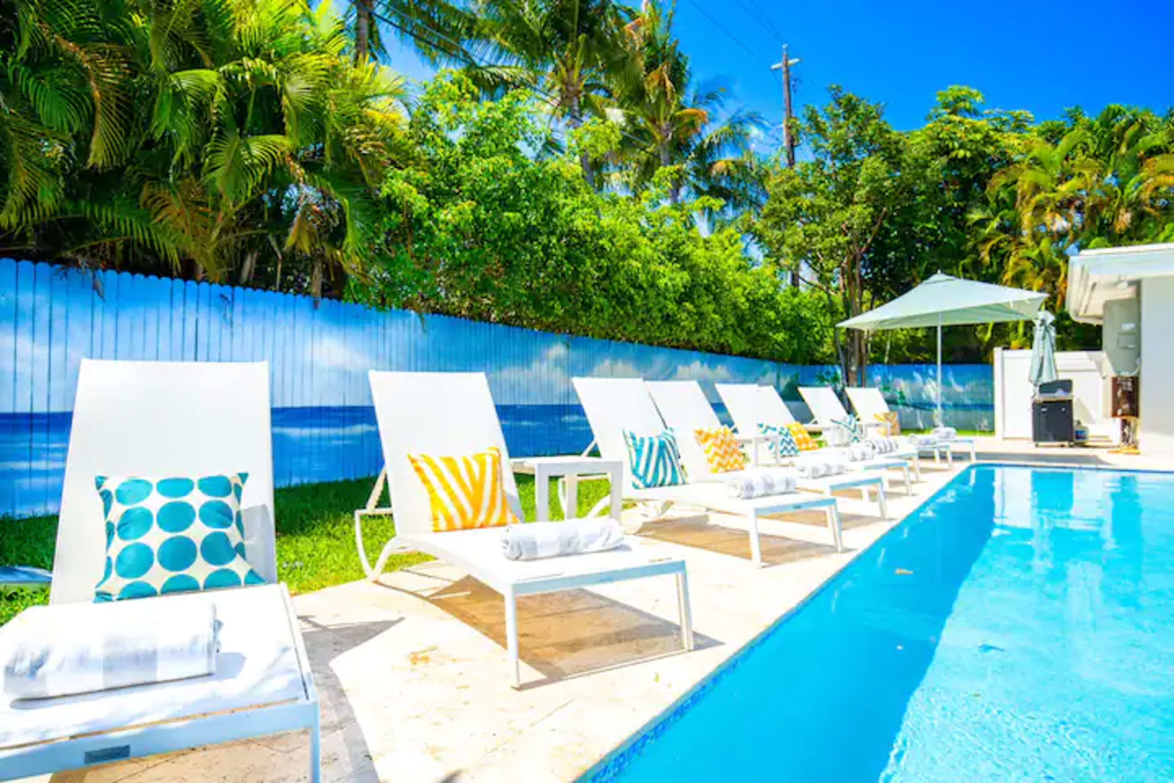 Lounge chairs with colorful cushions line a swimming pool surrounded by lush greenery and palm trees.