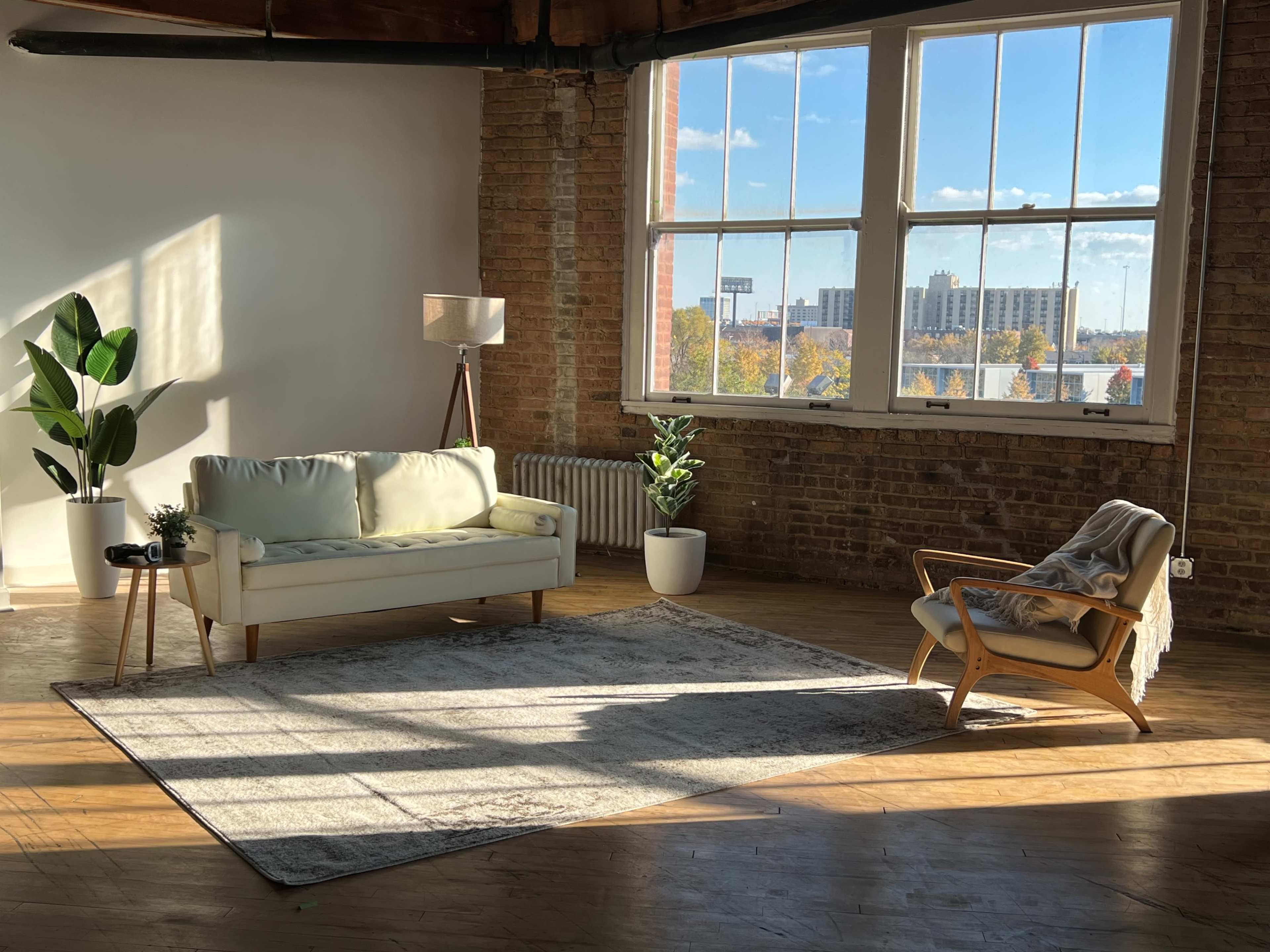 A modern living room features a light-colored sofa, a wooden chair, and indoor plants, illuminated by sunlight streaming through large windows.
