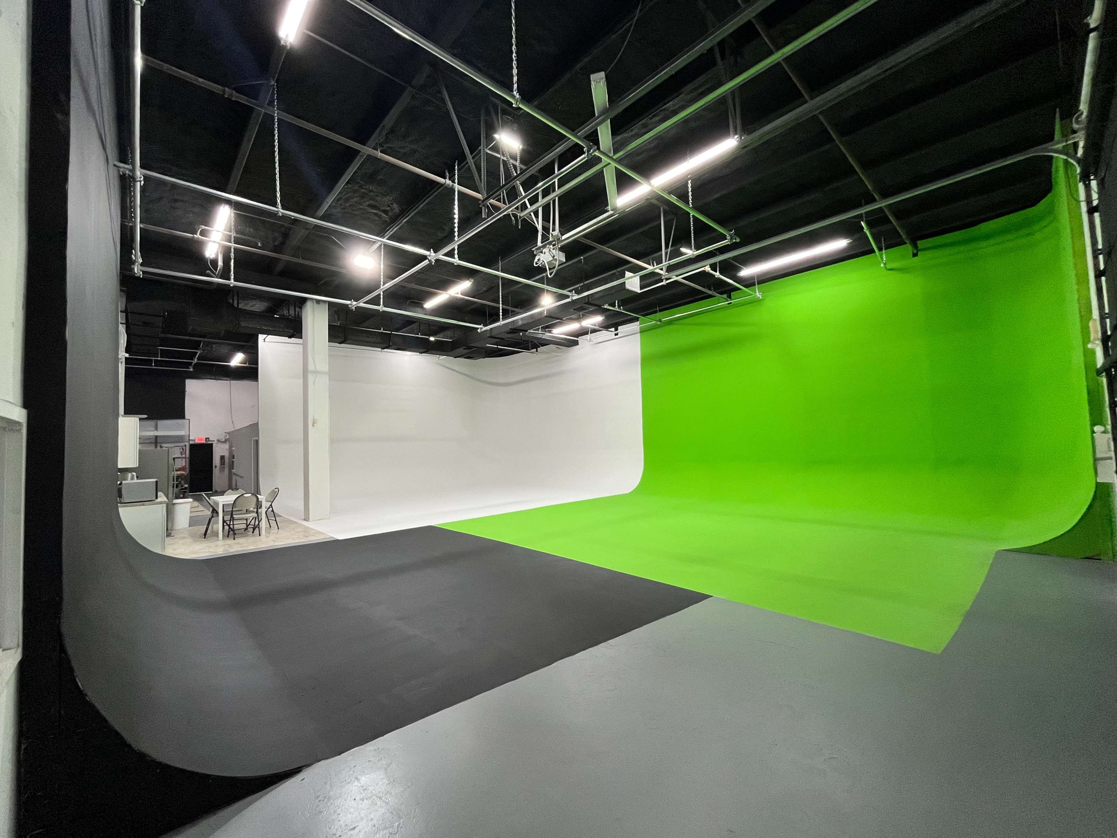 The image shows a spacious studio featuring a green backdrop on one side and a white backdrop on the other, with a black floor and some furniture in the background.
