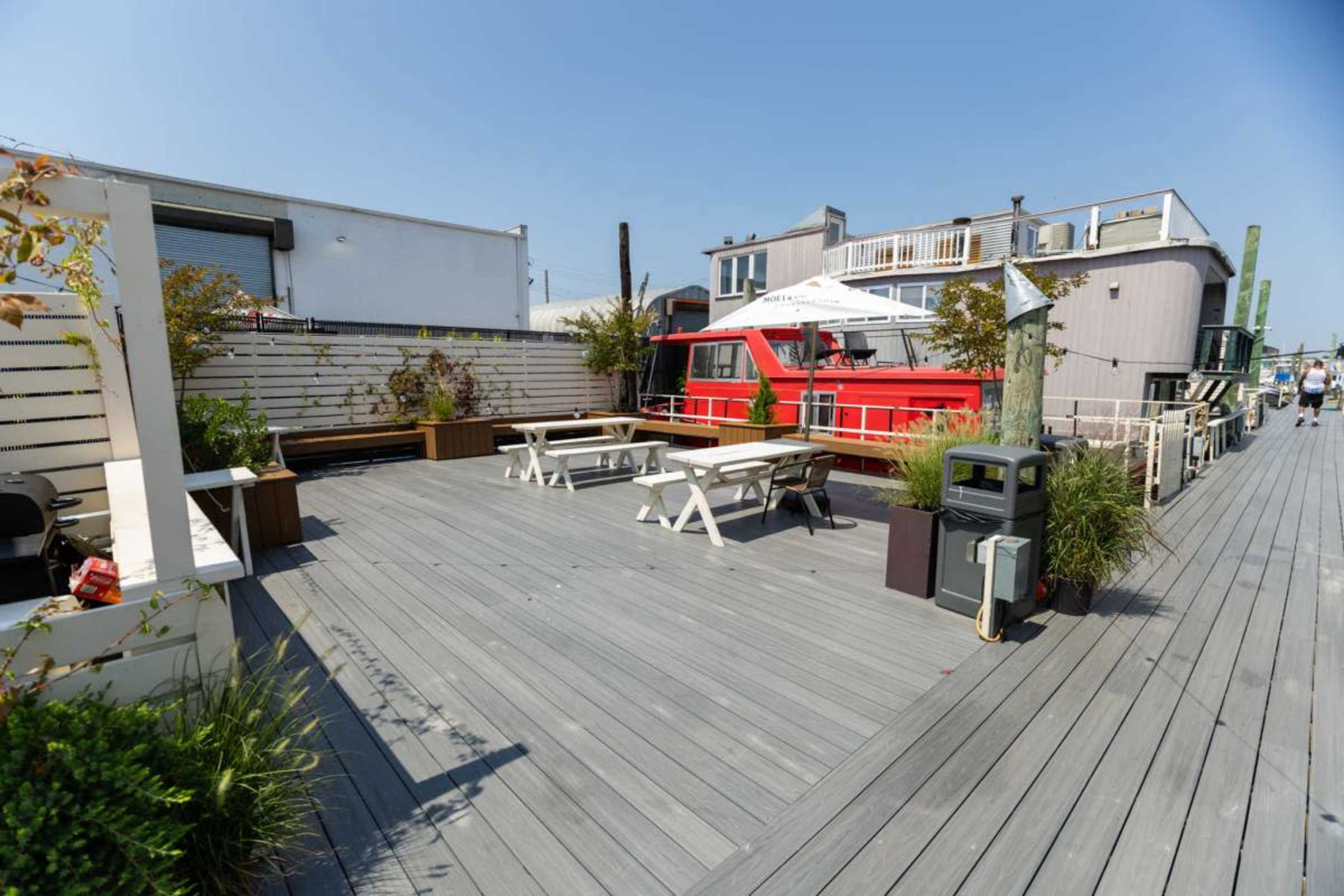 The image shows a wooden deck area with several tables and chairs, bordered by potted plants, alongside a red houseboat.