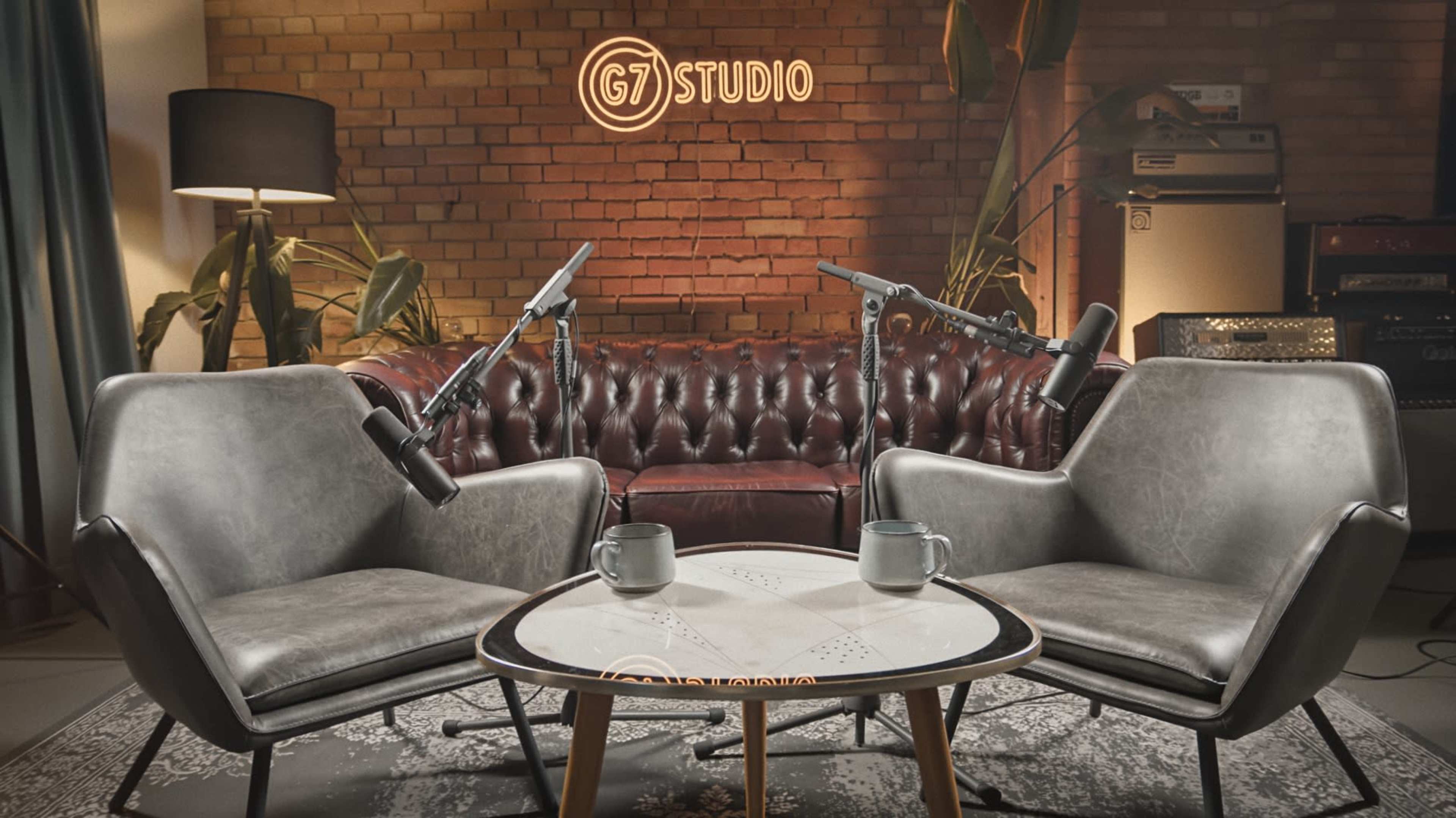 The image shows a cozy studio setup with two modern chairs, a small table between them, and microphones positioned in front of each chair, set against a brick wall with the text "G7 STUDIO" illuminated above.