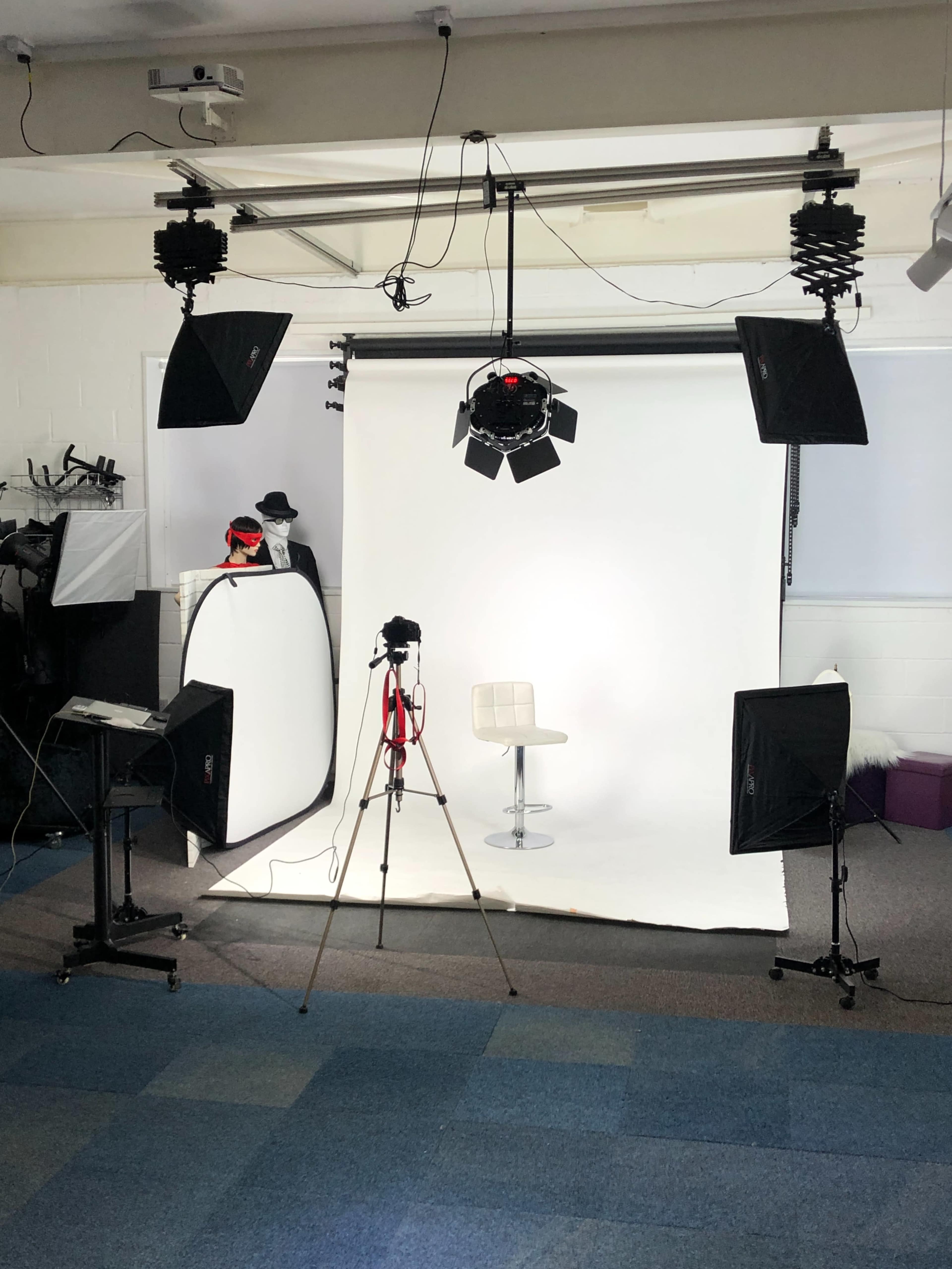 The image shows a photography studio setup featuring a white backdrop, a chair, lighting equipment, a camera on a tripod, and various props in the background.