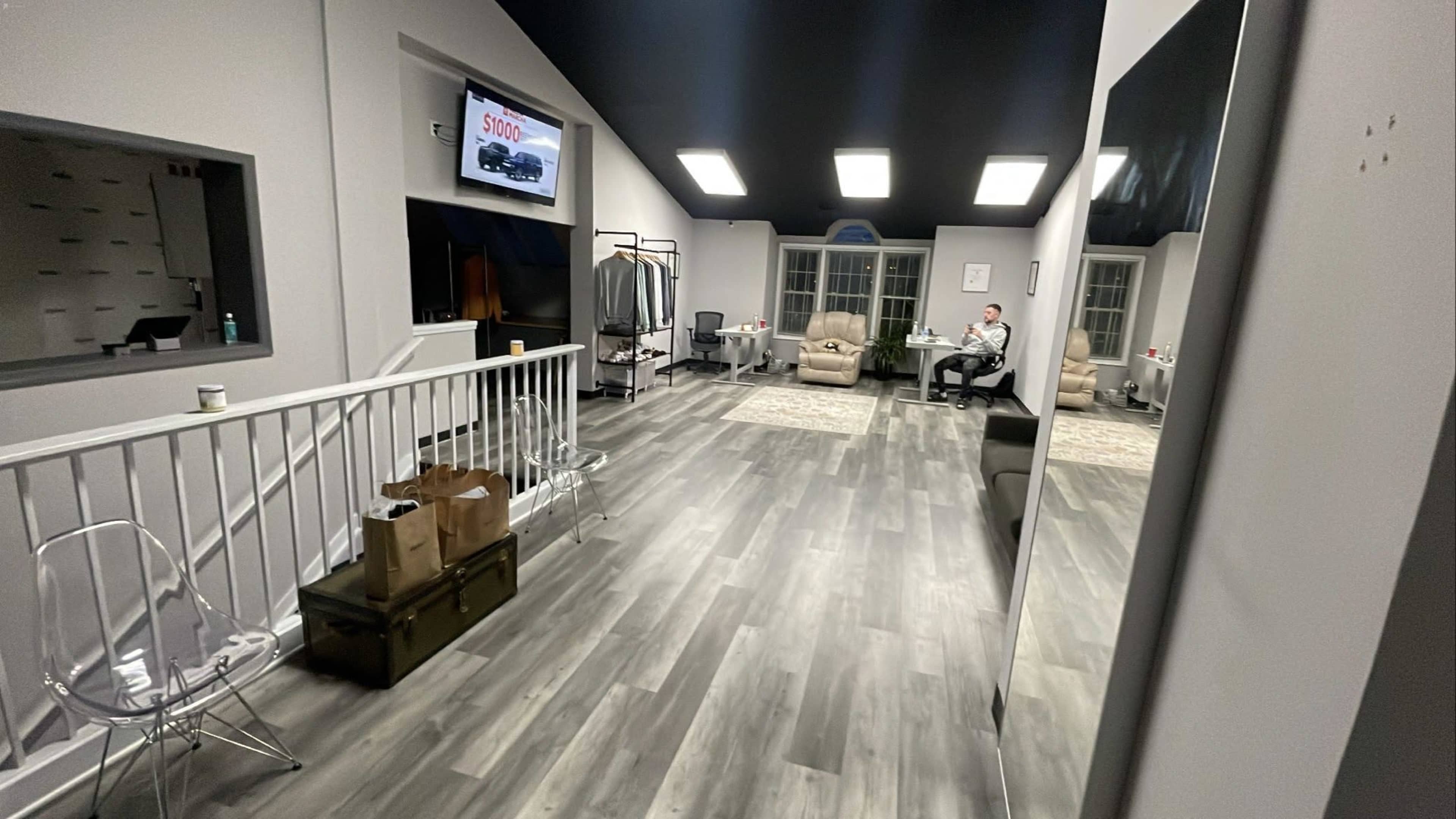 A spacious, modern room with gray wood flooring, a television on the wall displaying an advertisement, and a man seated on a couch surrounded by a few pieces of furniture and clothing racks.