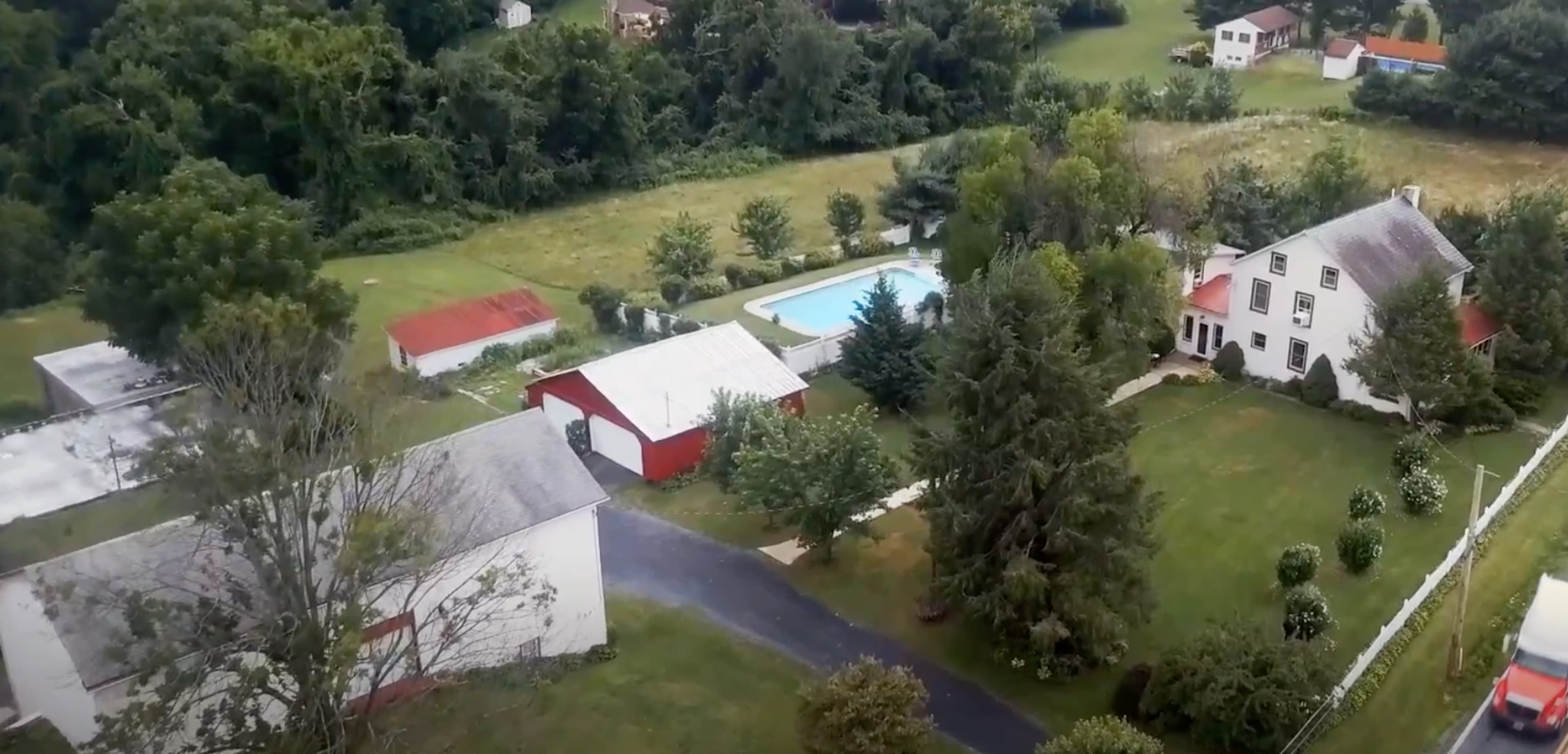 The image shows an aerial view of a rural property featuring a house, a swimming pool, several outbuildings, and landscaped grounds.