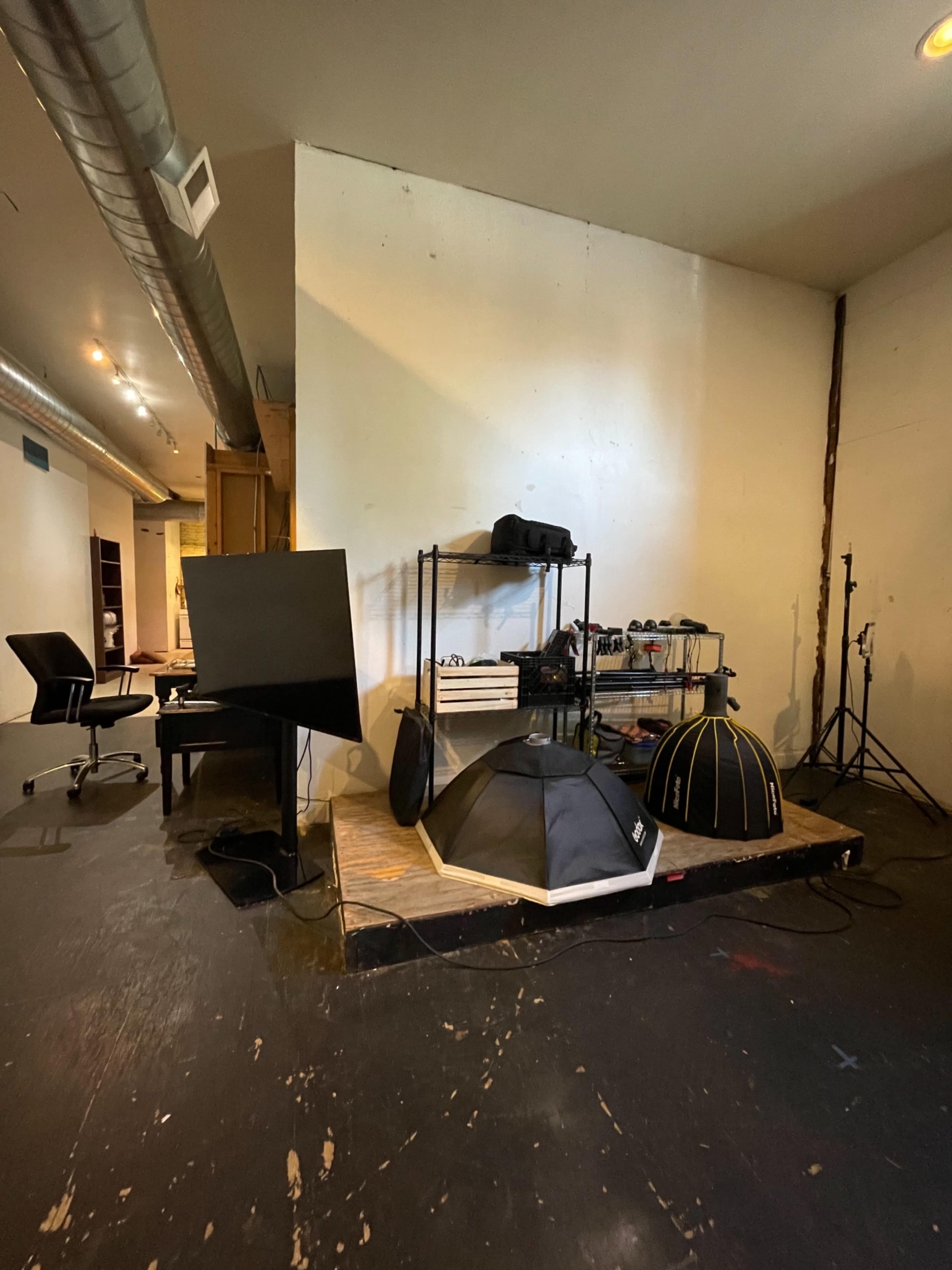 The image shows a workspace featuring a large monitor, a rolling shelf with equipment, and several items including tents arranged on a wooden platform in a sparsely furnished room.