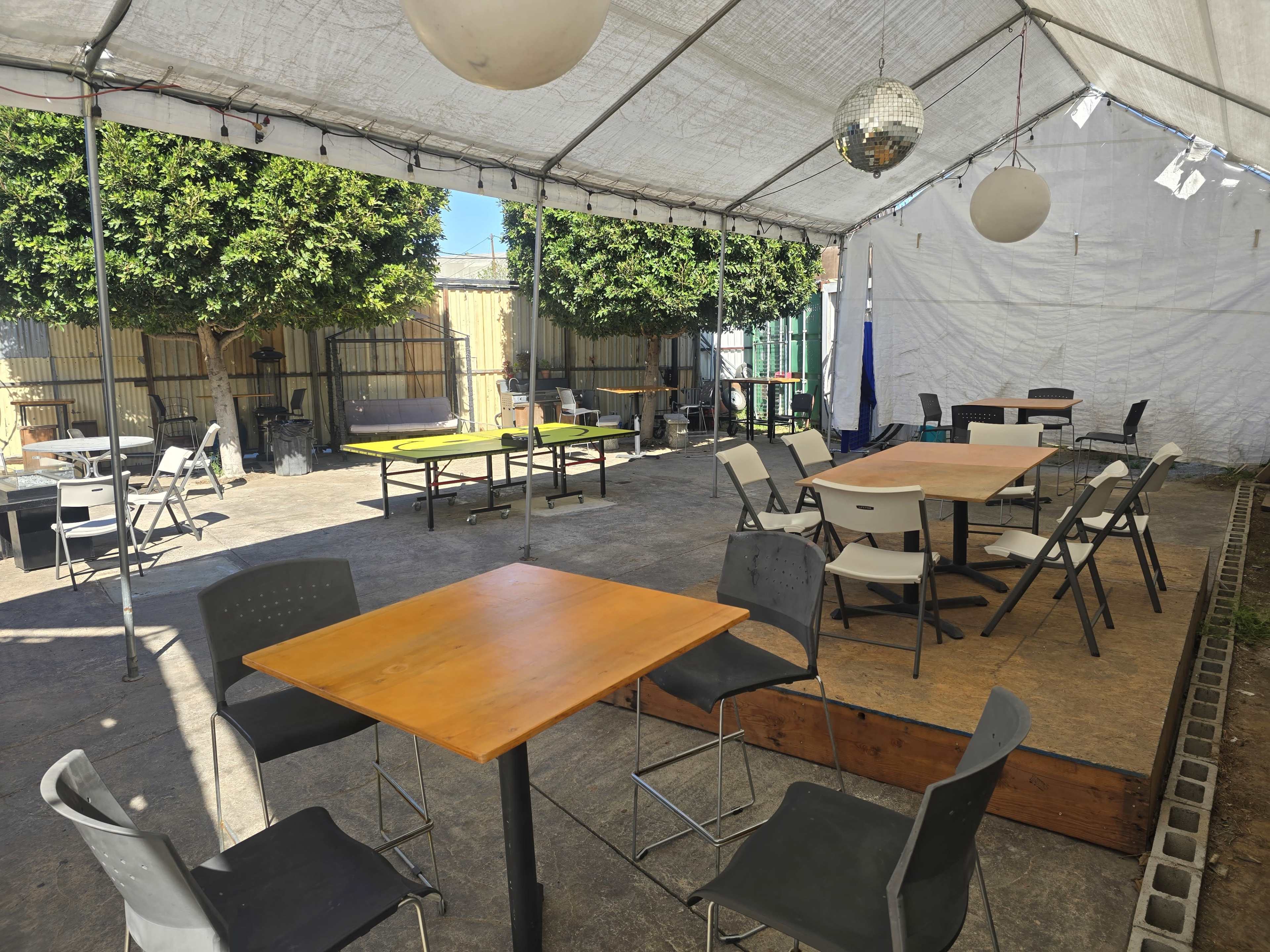 The image shows an outdoor seating area with several tables and chairs under a large white canopy, surrounded by greenery.