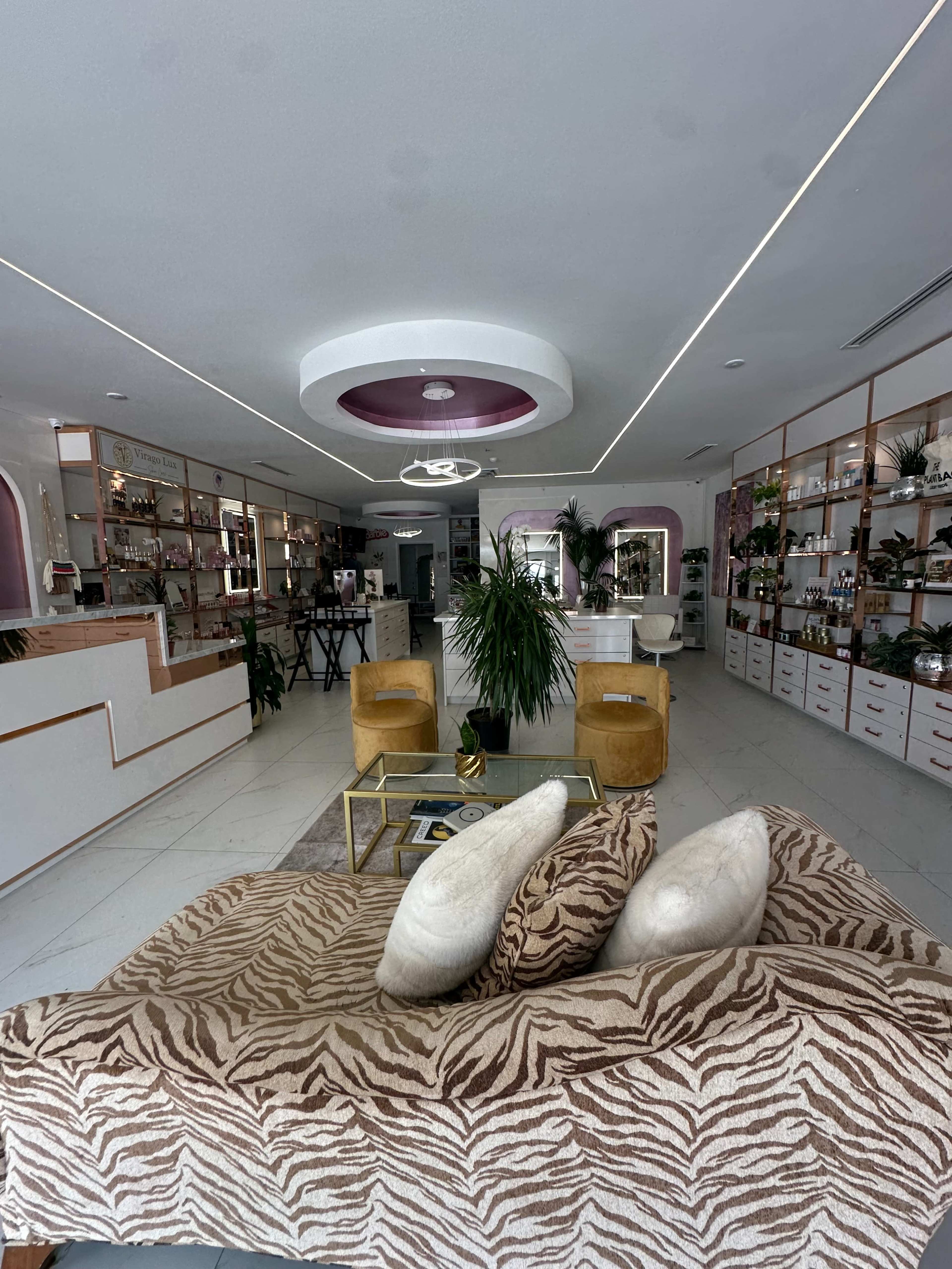 The image shows a modern interior of a beauty or wellness shop featuring a stylish seating area, a reception desk, and shelves filled with products.