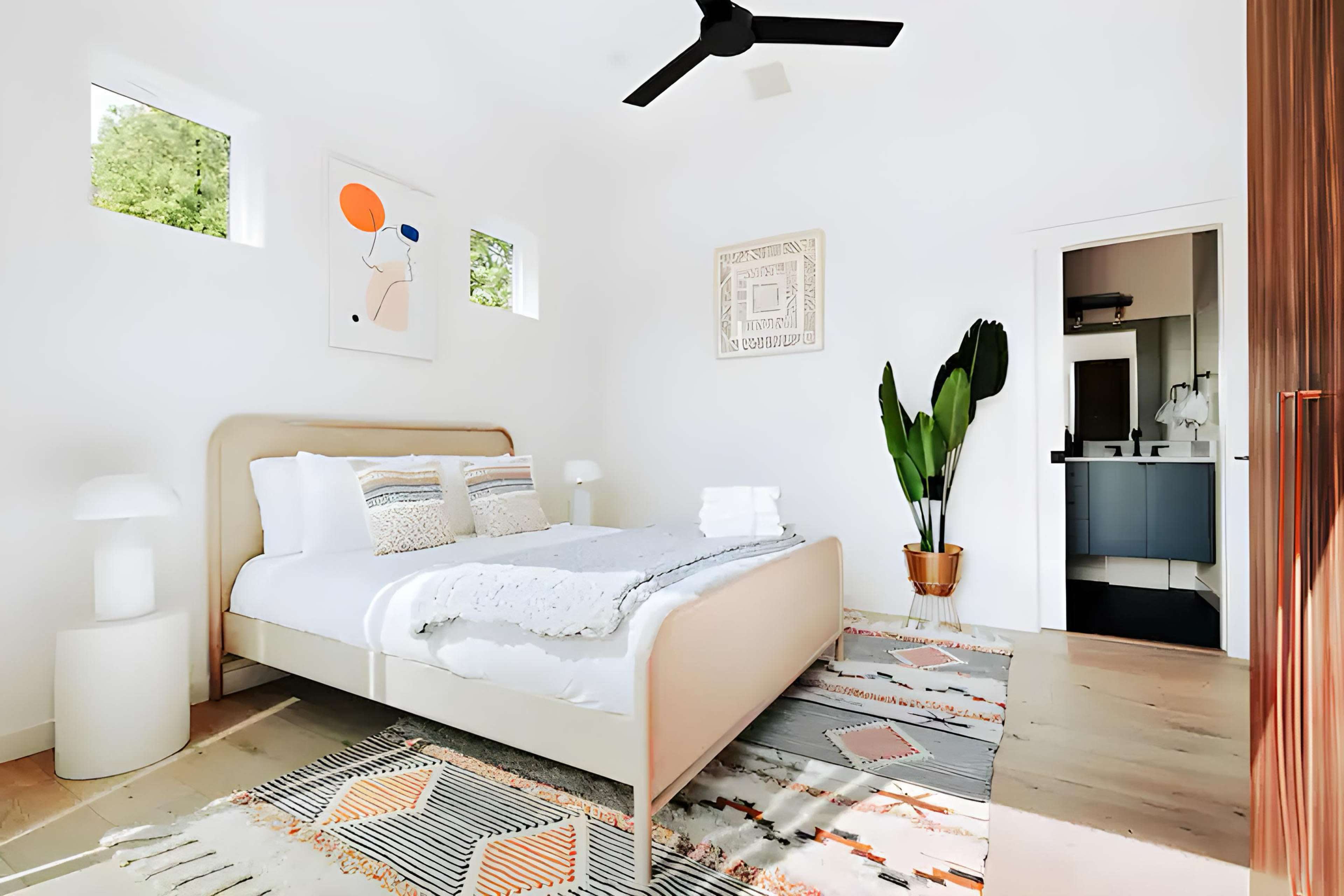 The image shows a bright bedroom with a bed, decorative pillows, a patterned rug, and a potted plant beside the door to a bathroom.